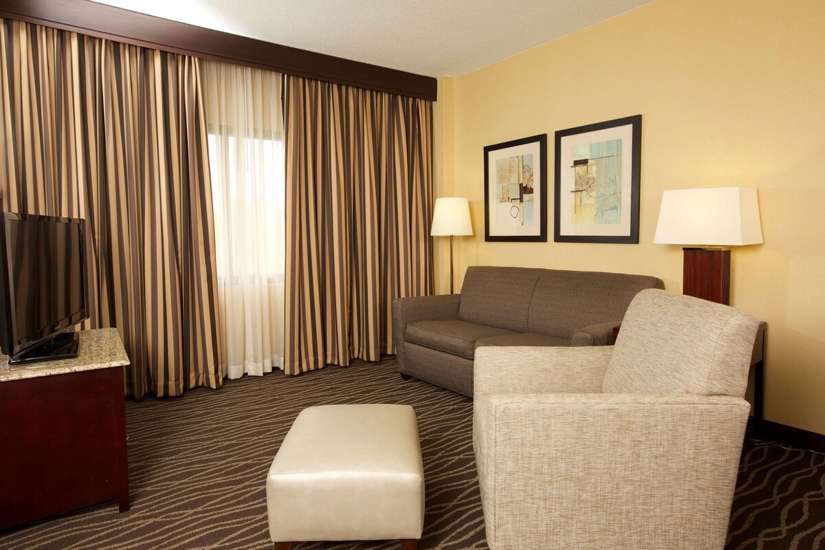 A hotel room with a sofa, armchair, ottoman, TV, lamps, and two framed pictures on the wall.
