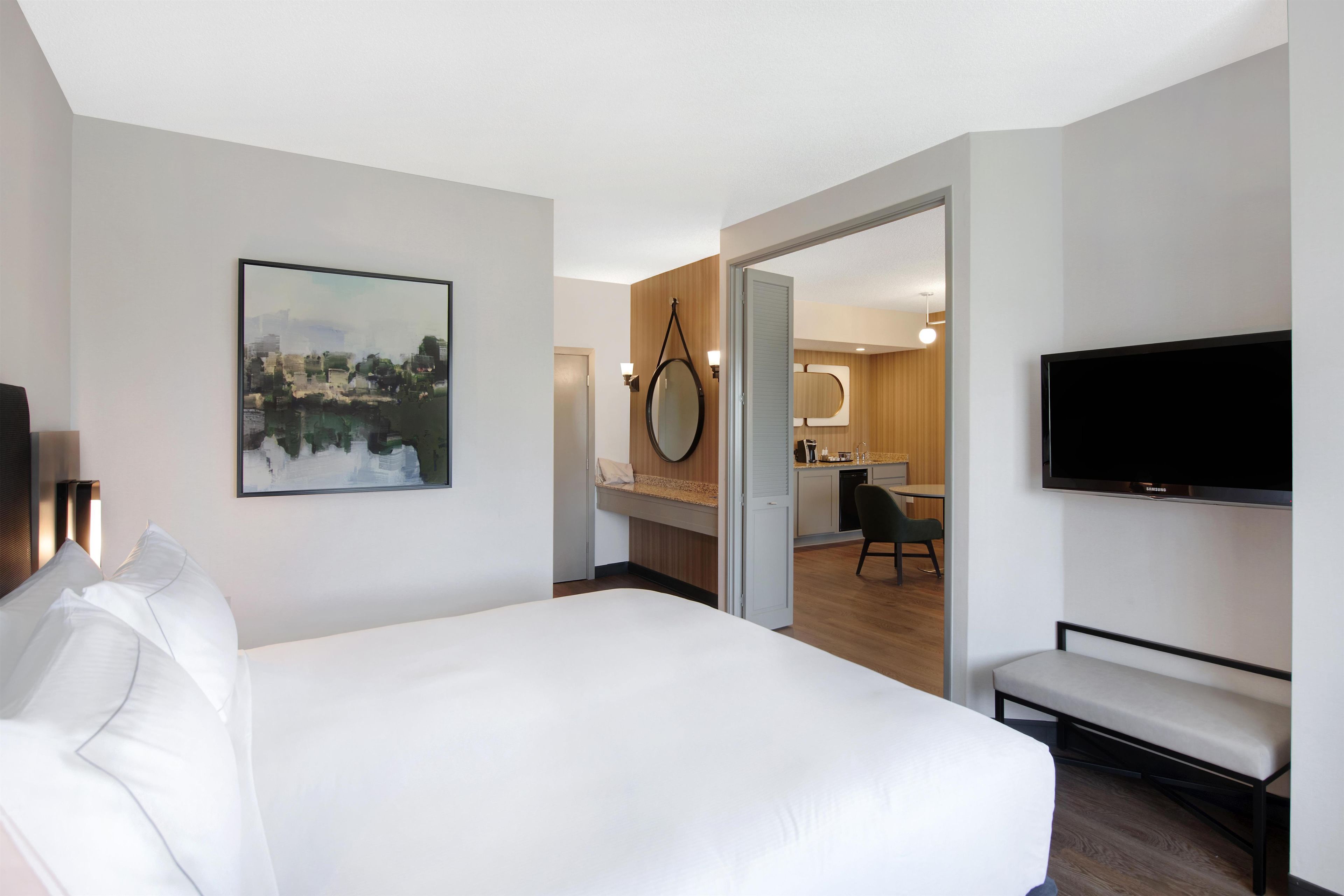 Une chambre d’hôtel avec un grand lit, une télévision, un tableau au mur et une vue sur la cuisine.