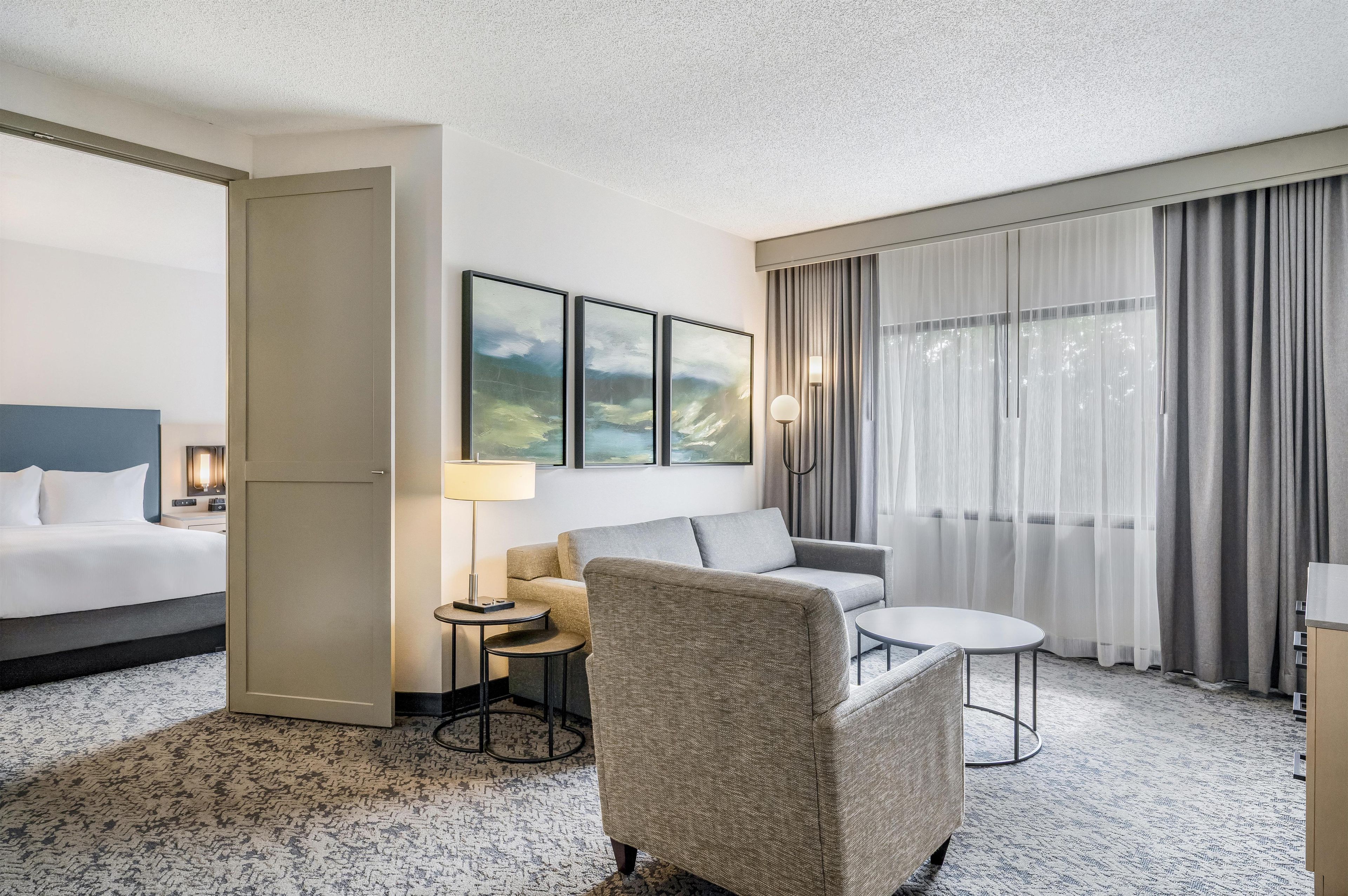 A hotel room with a bed, a sofa, a chair, a small table, and a lamp. There are three framed pictures on the wall.