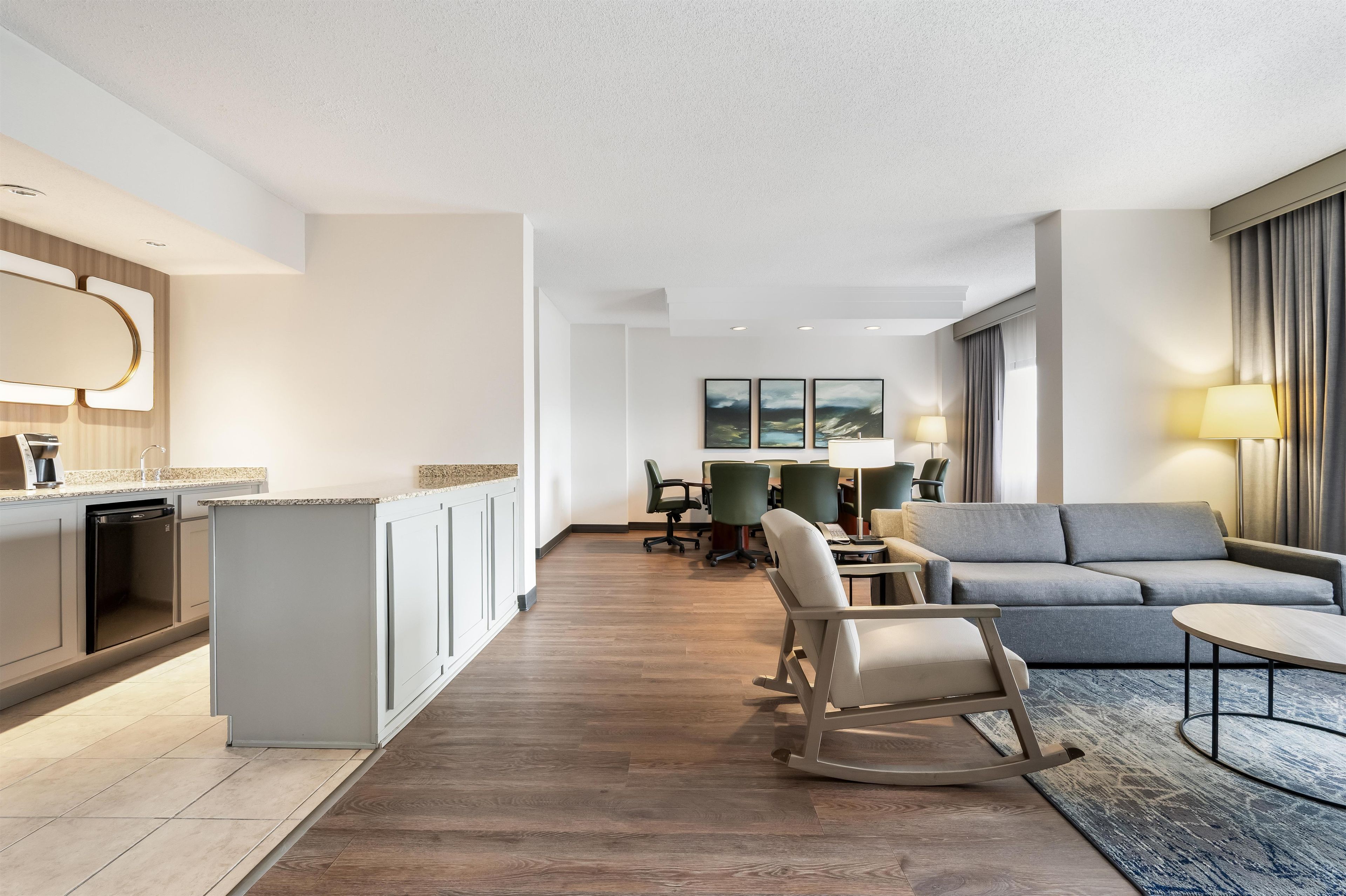 A modern hotel room with a kitchenette, a dining area, a living room with a sofa and a coffee table, and a large window with curtains.