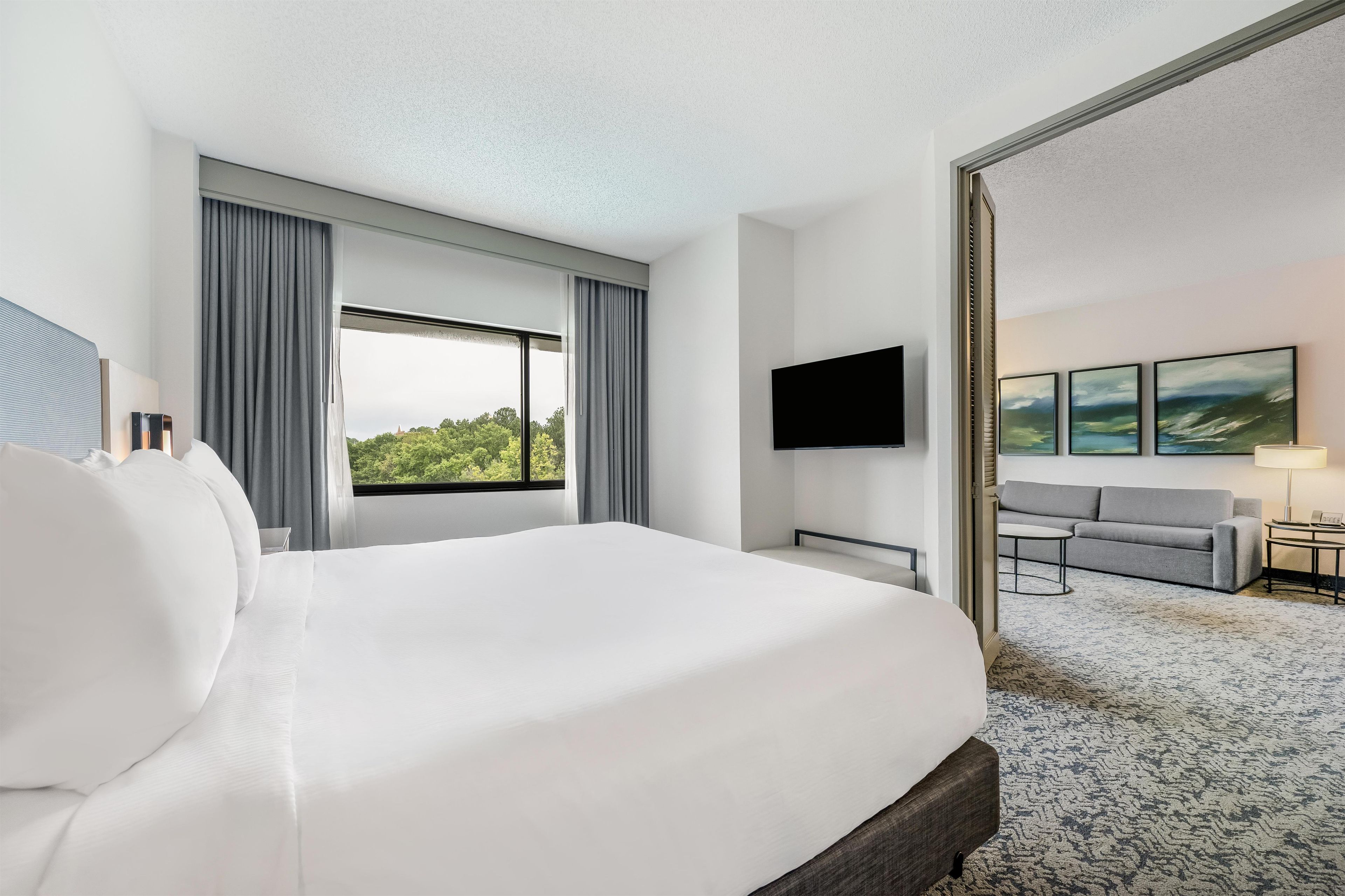 Una habitación de hotel con una cama grande, un televisor de pantalla plana montado en la pared, un sofá y una mesita de noche con una lámpara.