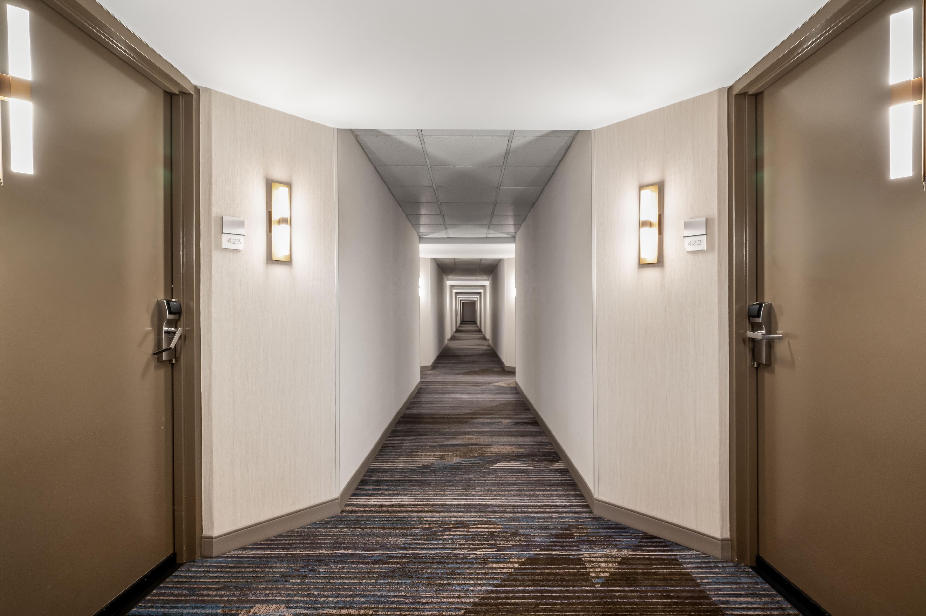 A hotel hallway with carpeted floors, doors on both sides, and illuminated wall sconces.