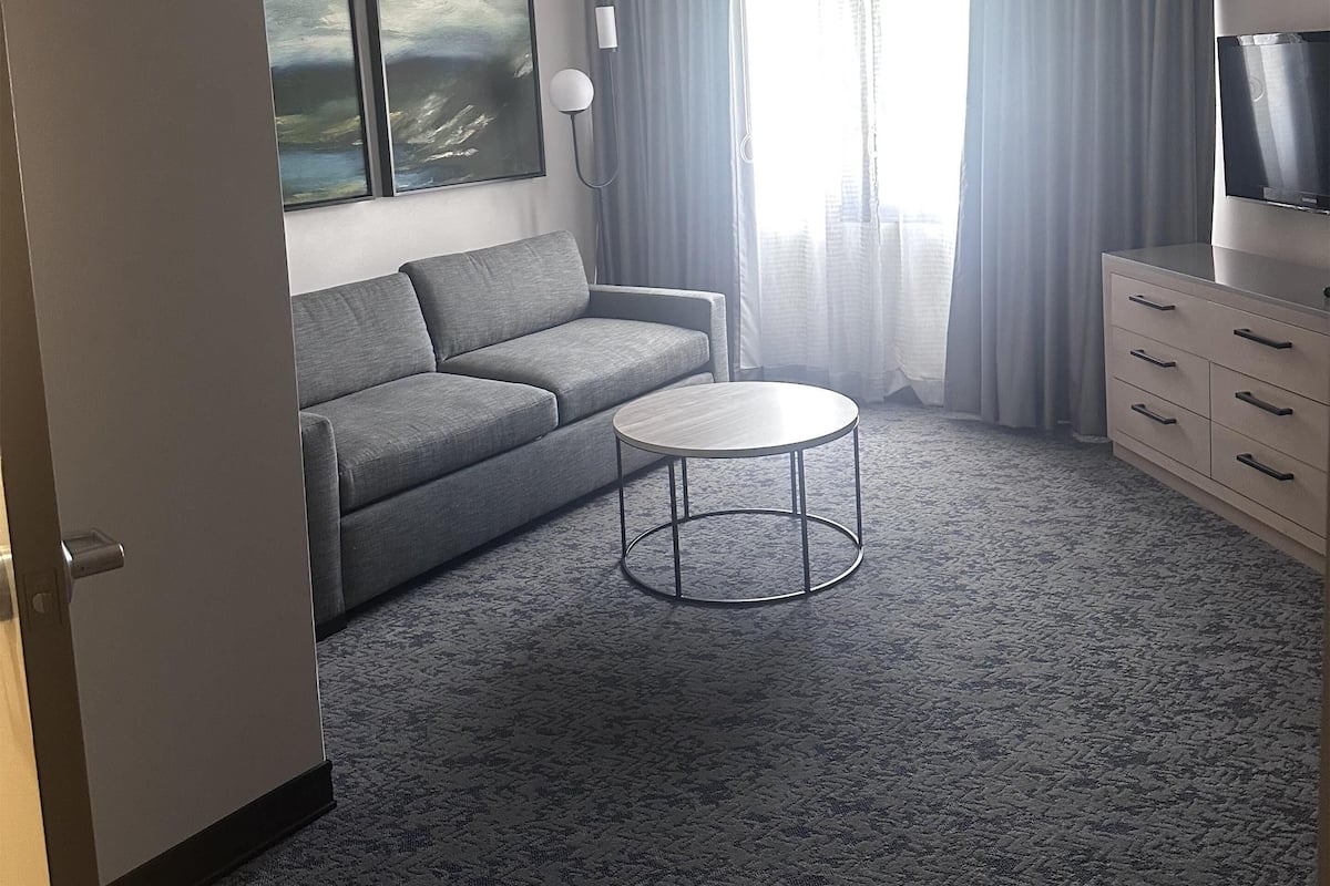 A modern hotel room with a grey sofa, a round coffee table, a TV on a stand, and a large window with curtains.