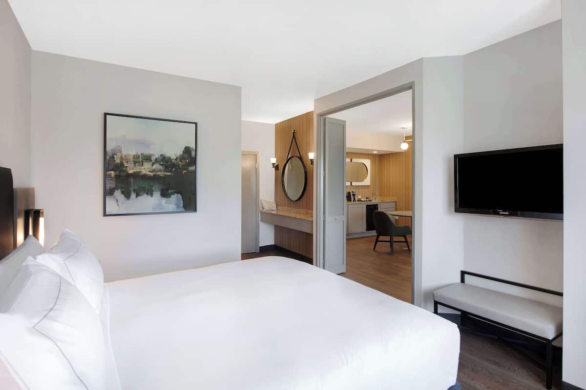 A hotel room with a bed, a TV, a painting, and a view into the kitchen area.