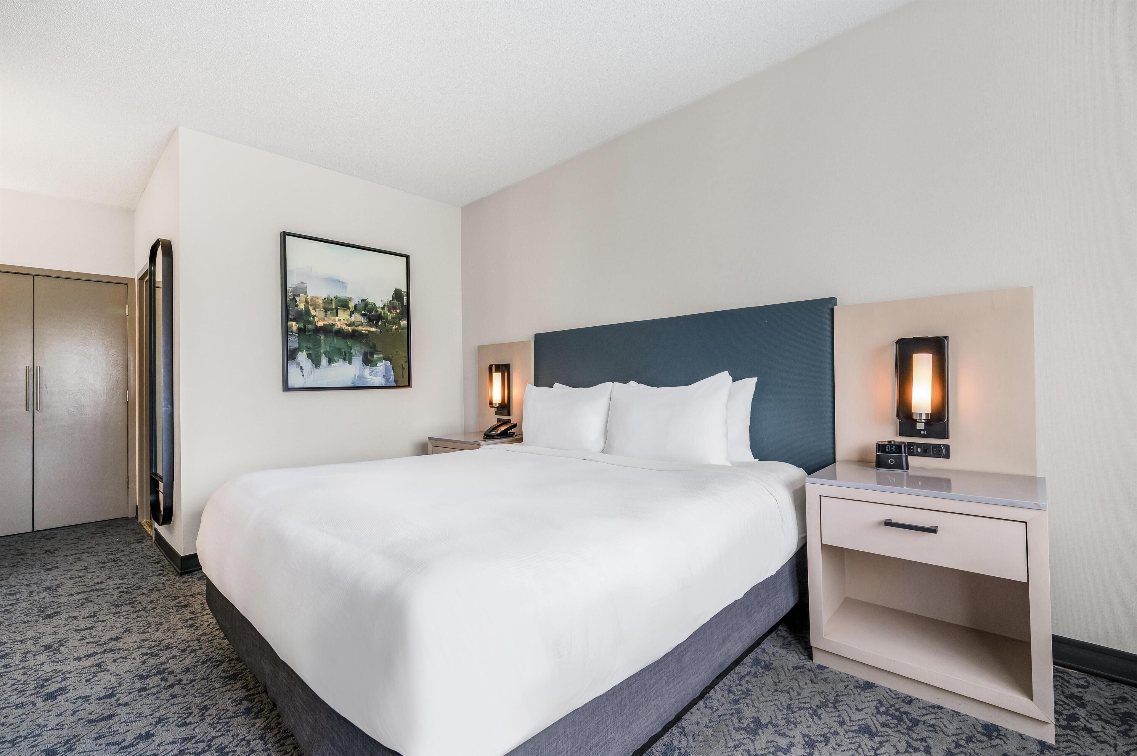 Een hotelkamer met een bed, nachtkastje, wandklok en een schilderij.