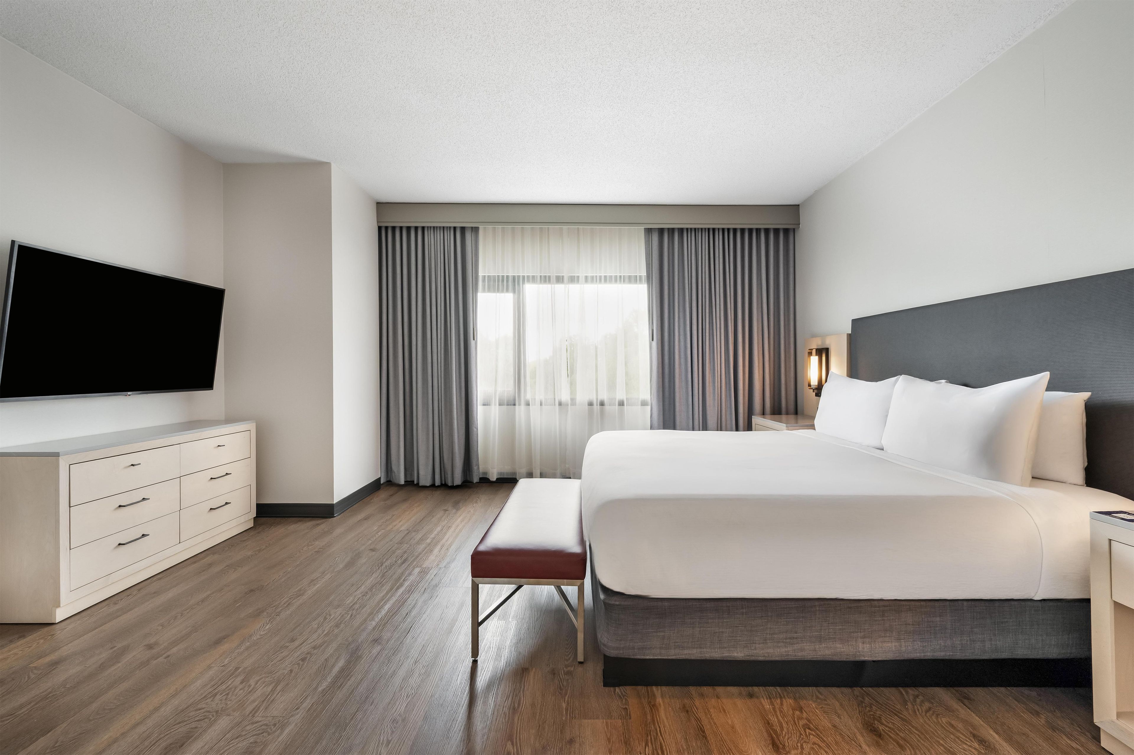 A hotel room with a bed, a flat-screen TV, a desk, and a window with curtains.