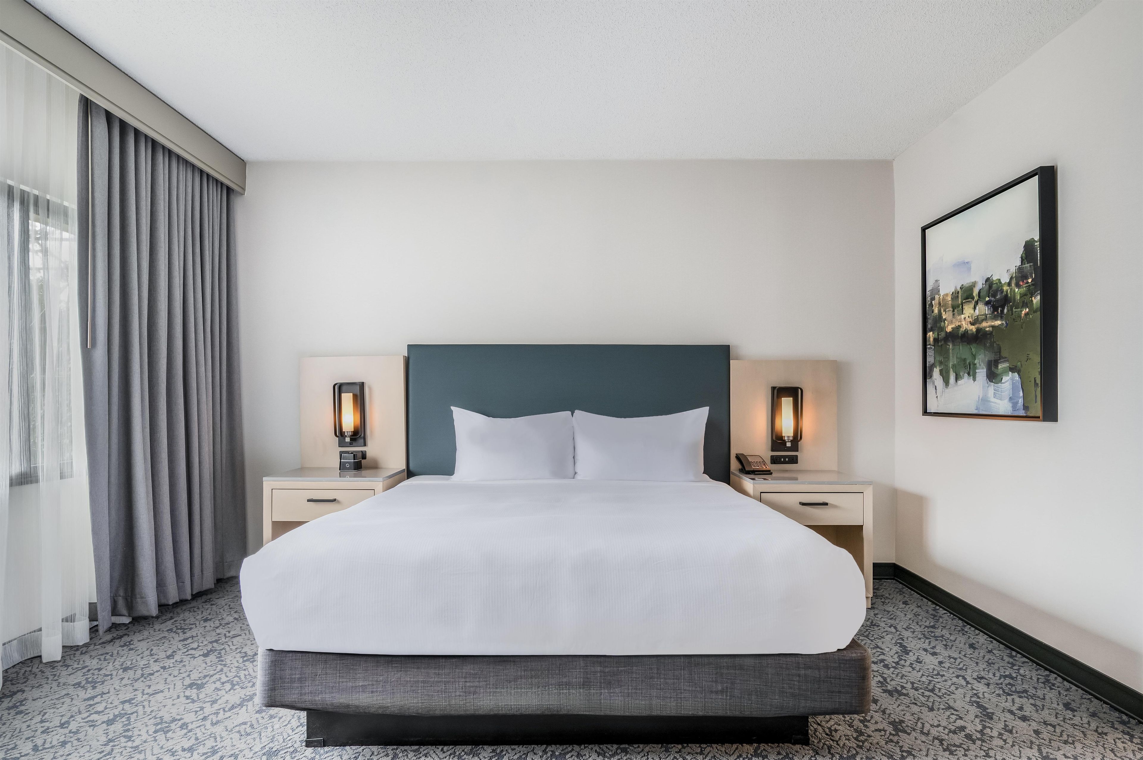 Une chambre d’hôtel avec un grand lit, deux tables de chevet avec des lampes, un tableau au mur et une fenêtre avec des rideaux.