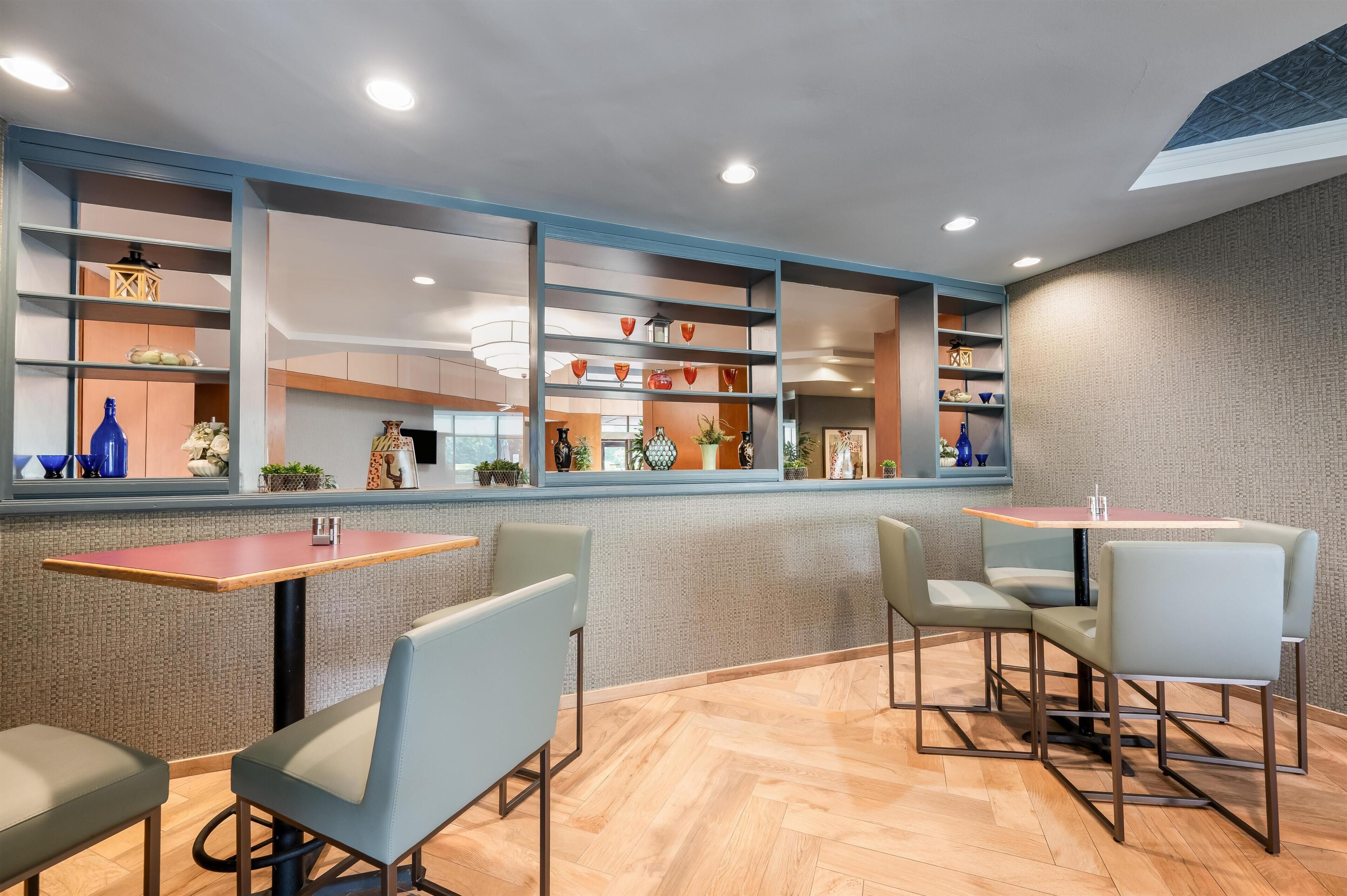 Un interior moderno con una zona de estar que incluye mesas y sillas, un estante decorativo con diversos objetos y una pared con diseño.