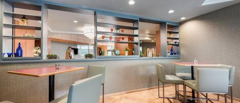 Un interior moderno con una zona de estar que incluye mesas y sillas, un estante decorativo con diversos objetos y una pared con diseño.
