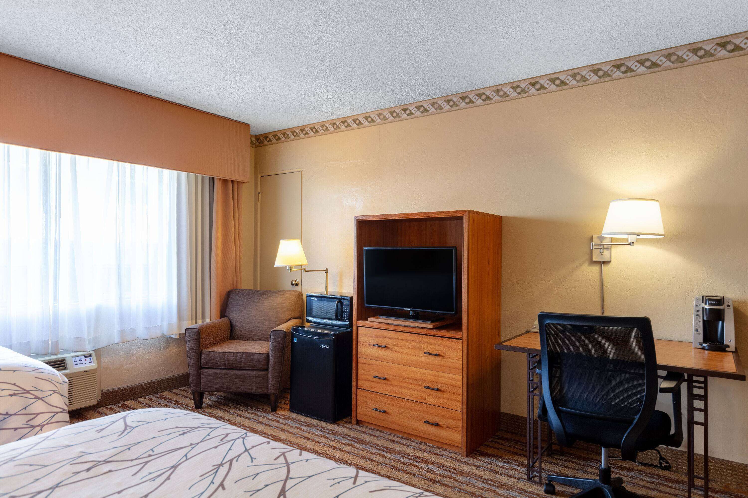 A hotel room with a bed, a desk with a computer, a TV, a chair, a microwave, and a coffee maker.