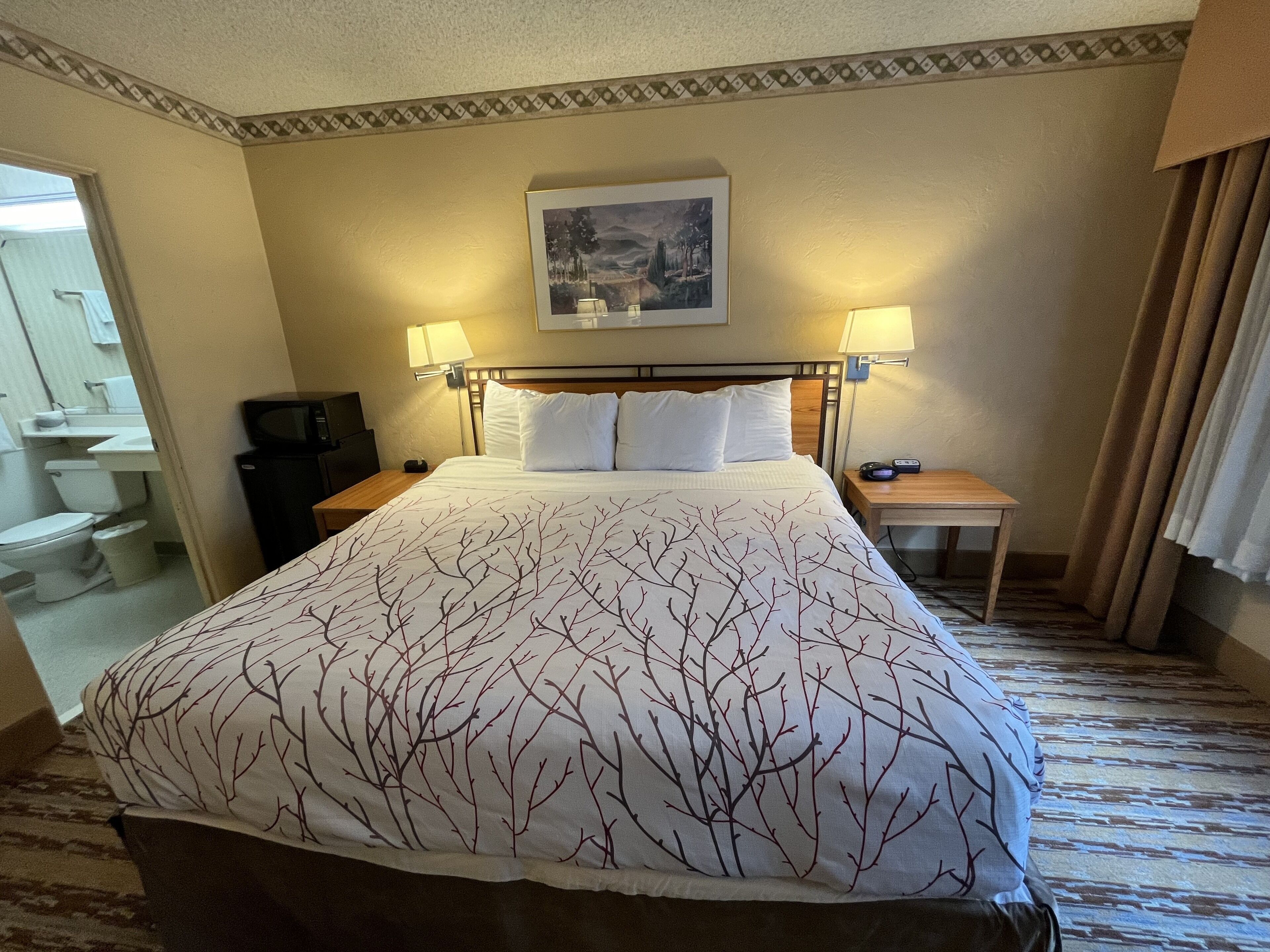 Habitación de hotel con una cama grande, dos lámparas de noche, una mesita de noche, un cuadro en la pared y un baño visible a través de una puerta abierta.