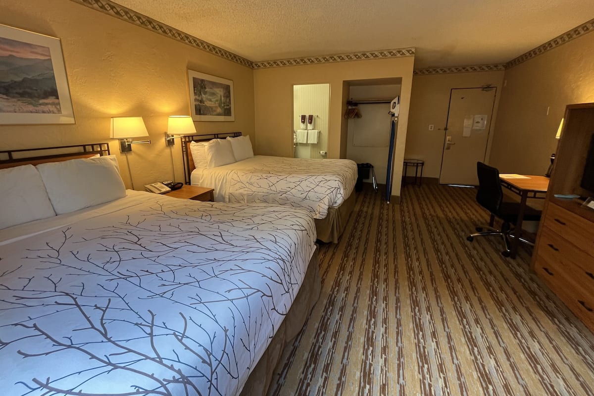 Quarto de hotel com duas camas, uma escrivaninha, uma cadeira, um abajur e um espelho.