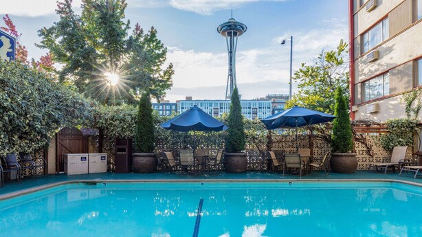 A swimming pool with lounge chairs and umbrellas, surrounded by greenery and a view of the Space Needle. - Travelodge by Wyndham Seattle By The Space Needle (Seattle)