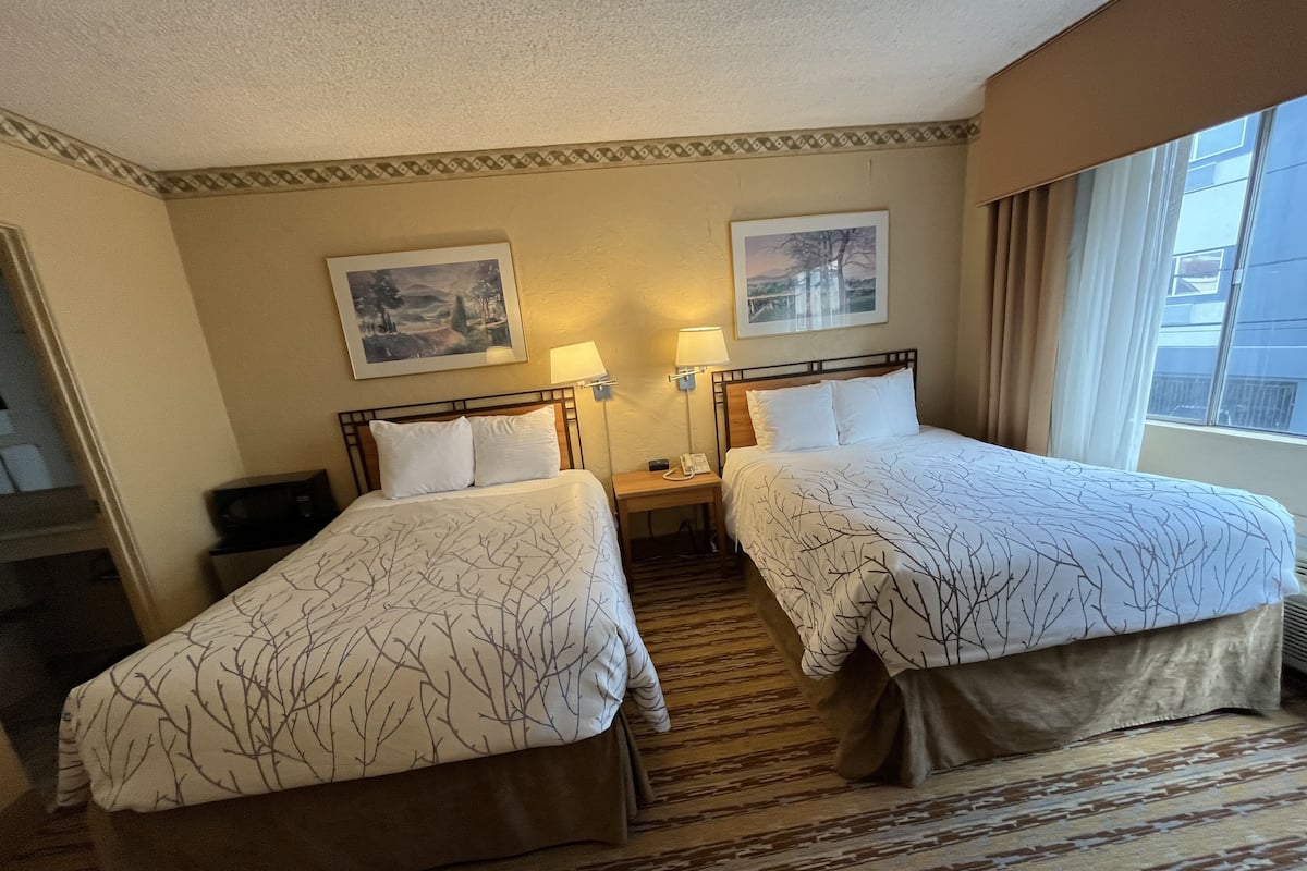 Quarto de hotel com duas camas de solteiro, uma mesa de cabeceira com abajur, telefone e duas fotos emolduradas na parede.