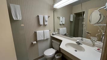 Un baño de hotel estándar con inodoro, lavamanos y espejo. Hay varias toallas colgadas en la pared y un cesto de basura.