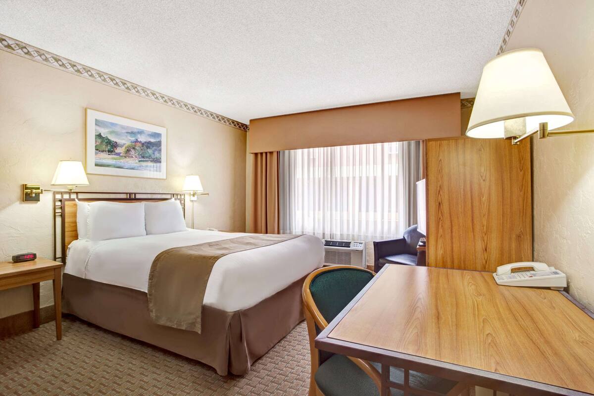 Quarto de hotel com cama, mesas de cabeceira, escrivaninha, cadeira, abajur, telefone e um quadro na parede.