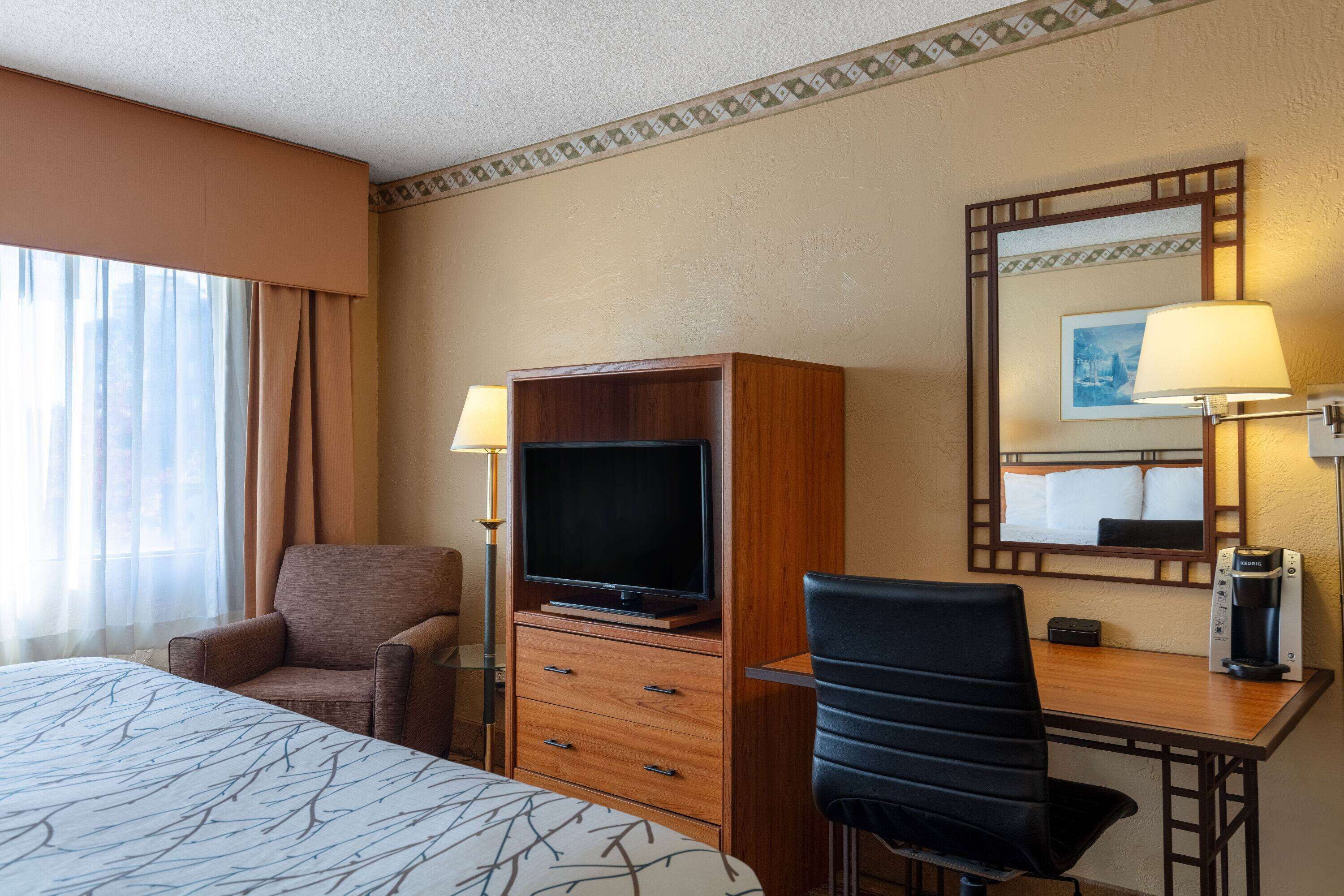 A hotel room with a bed, a TV, a desk, a chair, a coffee maker, and a lamp.