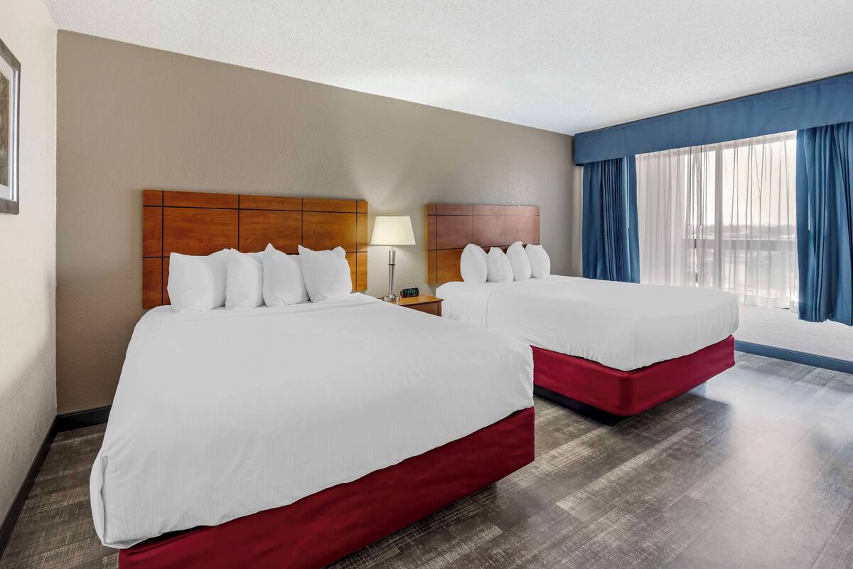 A hotel room with two beds, a nightstand with a lamp, and a large window with blue curtains.