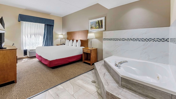 Une chambre d’hôtel avec un grand lit, une table de chevet en bois, un bureau avec une lampe, une salle de bain avec une baignoire et une fenêtre avec des rideaux.