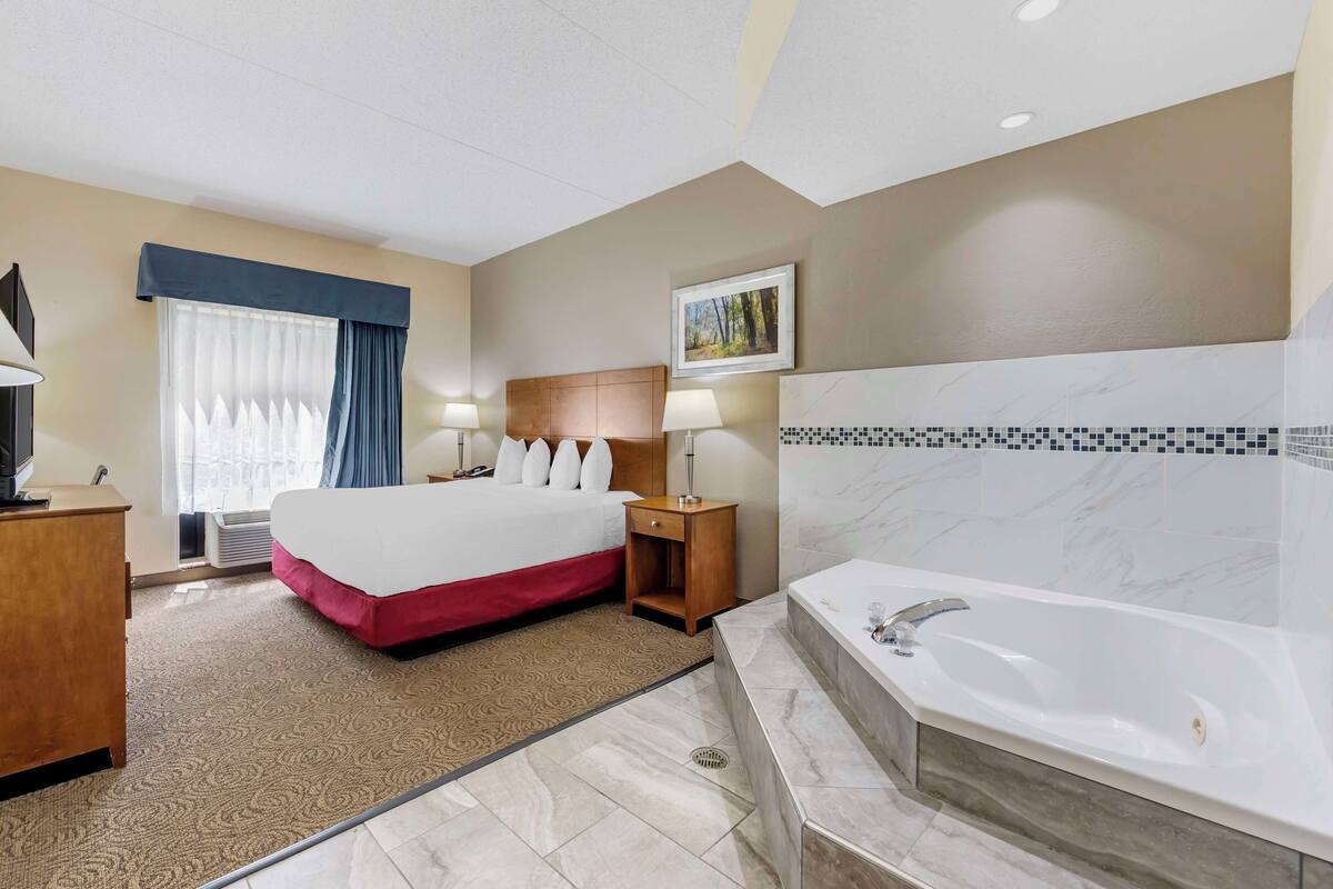A hotel room with a large bed, a wooden nightstand, a desk with a lamp, a bathroom with a bathtub, and a window with curtains.