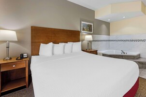 Habitación de hotel con una cama grande, un cabecero de madera, una mesita de noche con una lámpara, un teléfono y una pequeña mesa con una lámpara. Cuenta con un baño que incluye bañera y ducha.