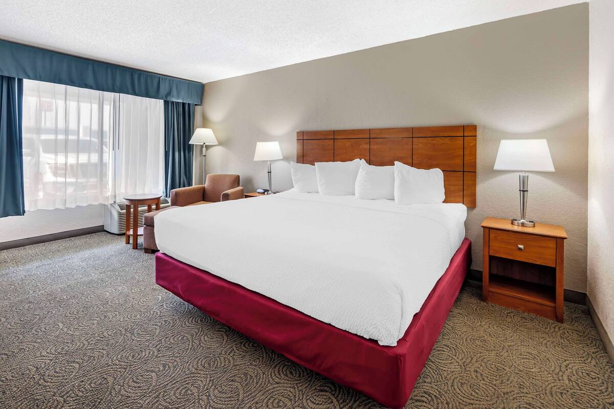 A hotel room with a large bed, two bedside tables with lamps, a chair, a desk, and a view of the outdoors through a window with blue curtains.
