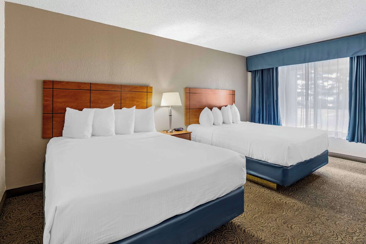 A hotel room with two beds, a nightstand, a lamp, and a large window with curtains.