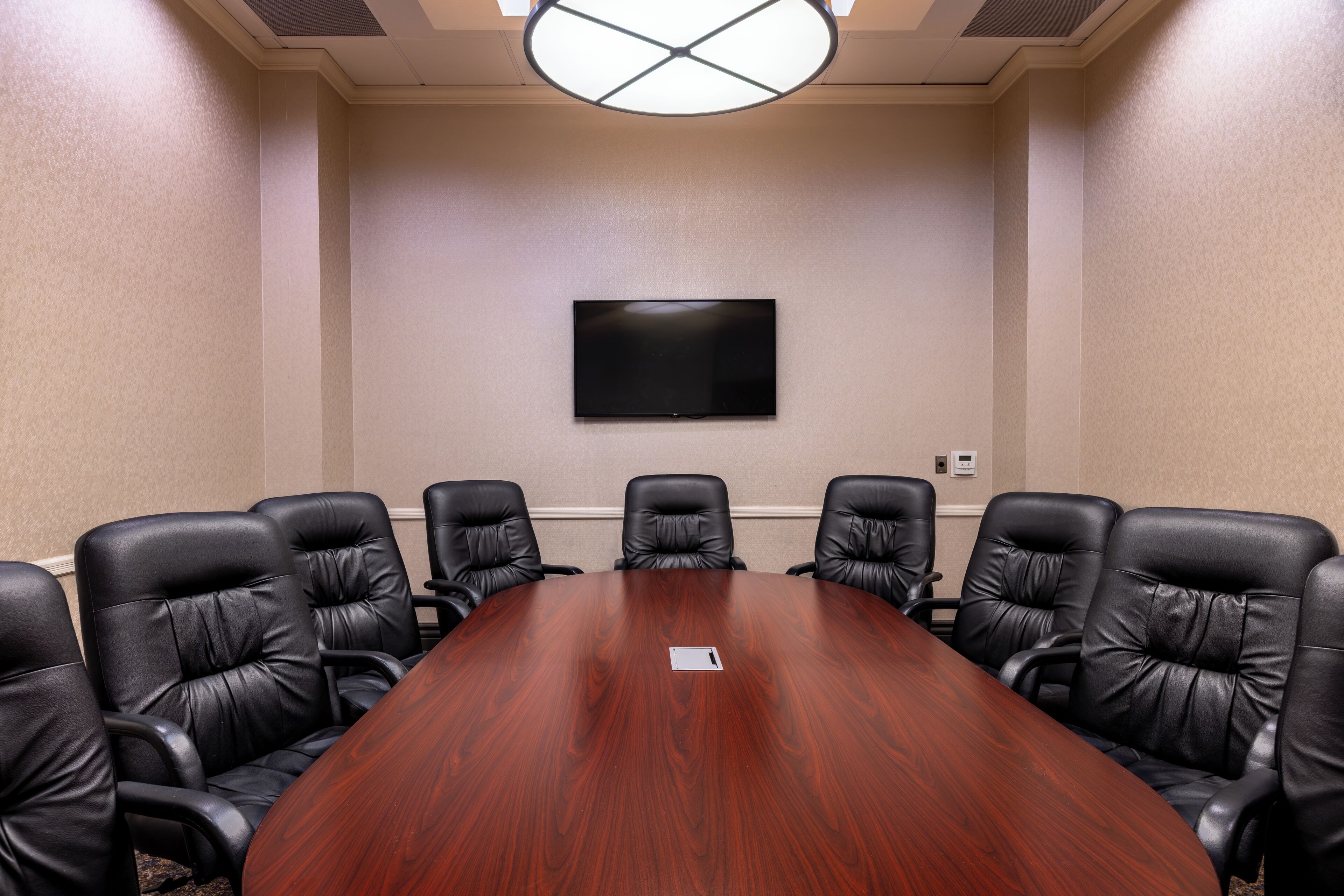 A conference room with a large wooden table, six black leather chairs, a flat-screen TV mounted on the wall, and a ceiling light fixture.