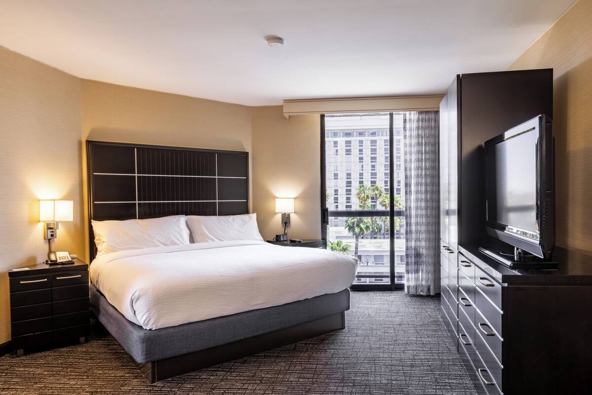Ein Hotelzimmer mit einem großen Bett, Nachttischlampen, einem Flachbildfernseher und Blick auf die Stadt durch ein großes Fenster.