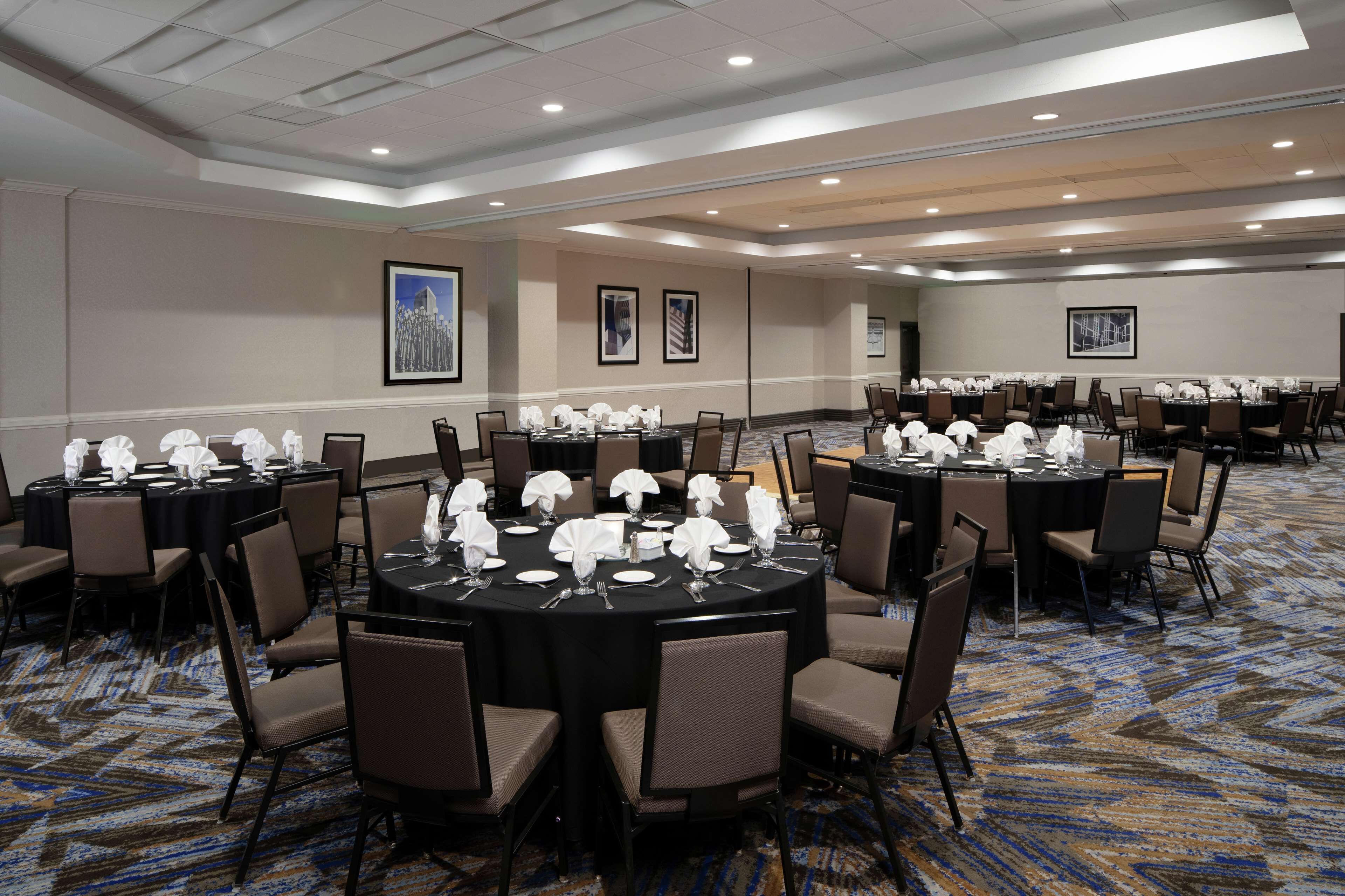 A conference room with multiple round tables, each set with white tablecloths, plates, and glassware. The room features a patterned carpet and beige walls with framed pictures.