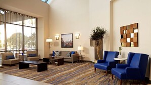 A spacious lobby with a high ceiling, large windows, a patterned carpet, a sofa set, a coffee table, a blue armchair, a small table with a lamp, and a decorative plant. - Embassy Suites by Hilton Los Angeles International Airport North (Los Angeles)