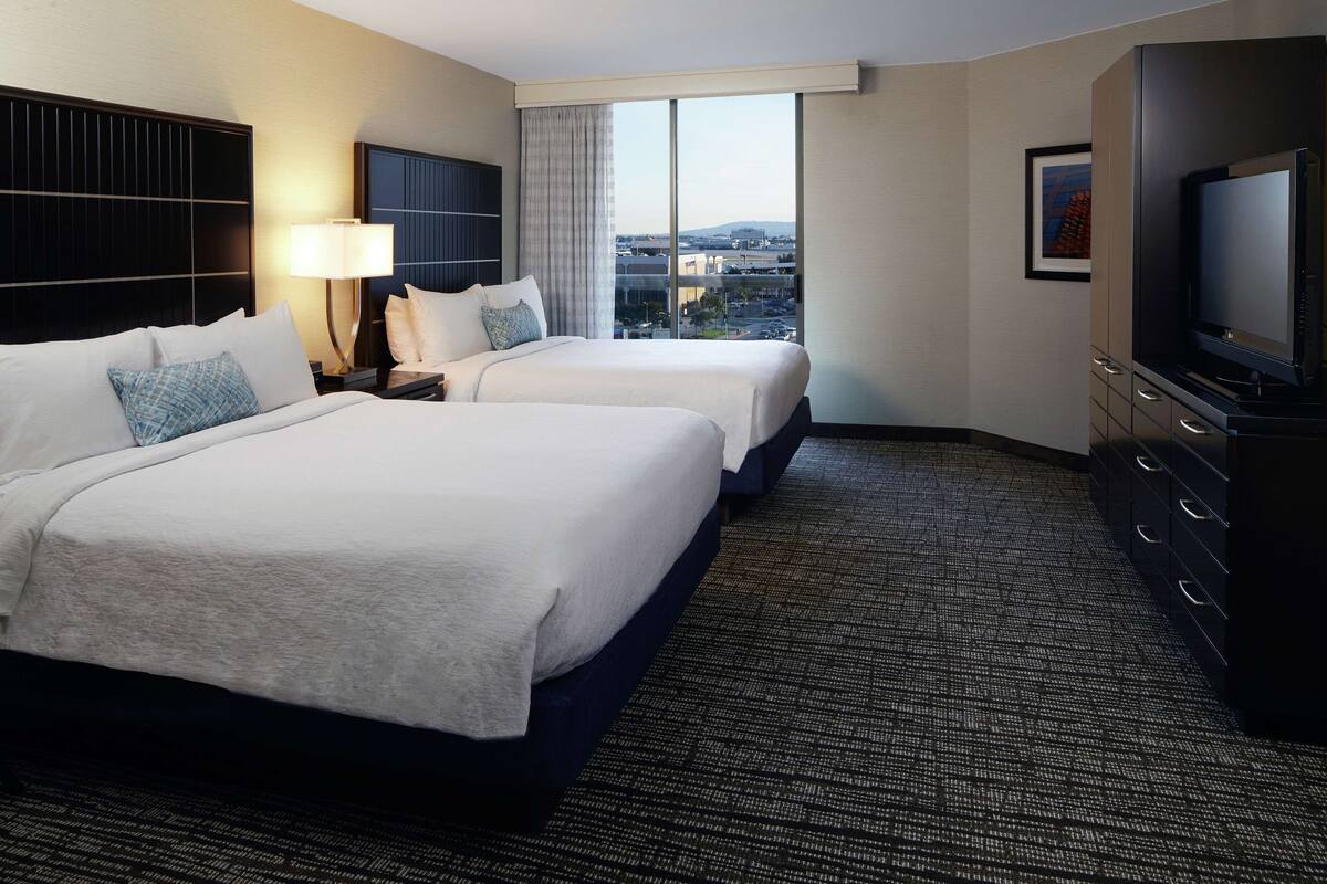 A hotel room with two beds, a TV, a desk, and a large window with a city view.