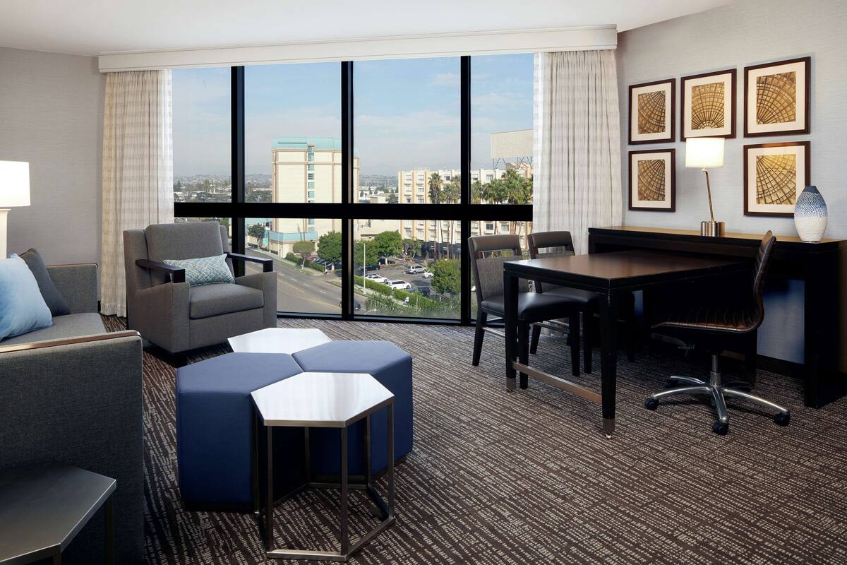 Ein Hotelzimmer mit großem Fenster, einer Sitzecke mit Tisch und gerahmten Kunstwerken an der Wand.