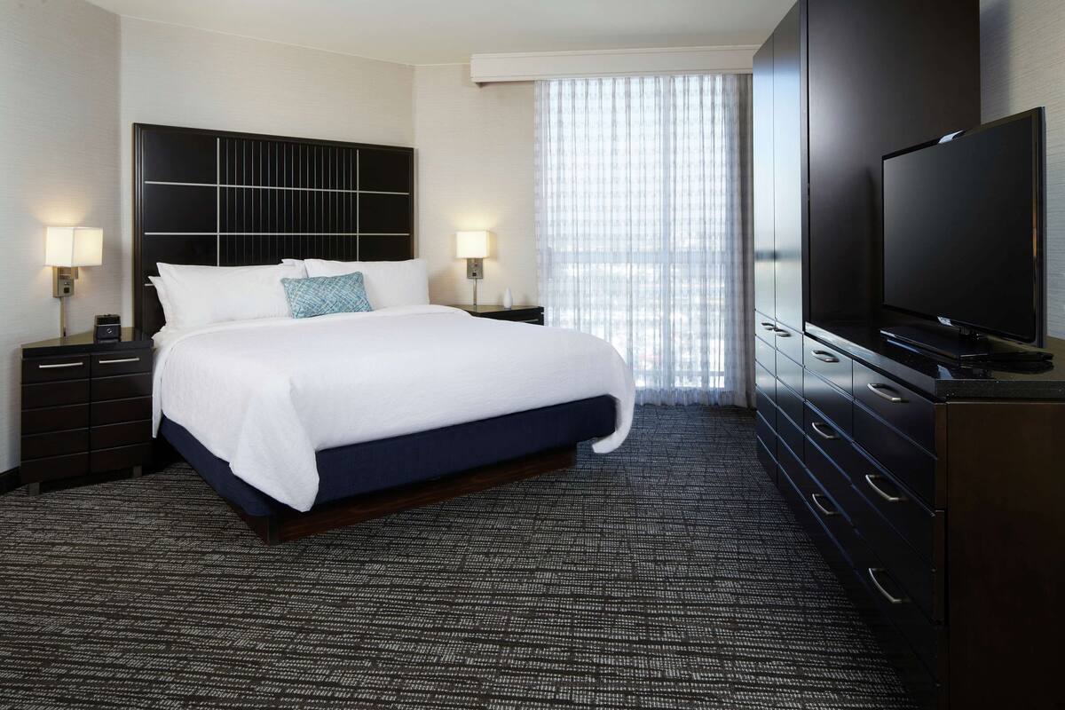 A hotel room with a large bed, a flat-screen TV, a dark wood dresser, and a large window with sheer curtains.