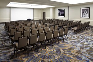 Un salón de conferencias con filas de sillas, una pantalla de proyector y cuadros en las paredes.