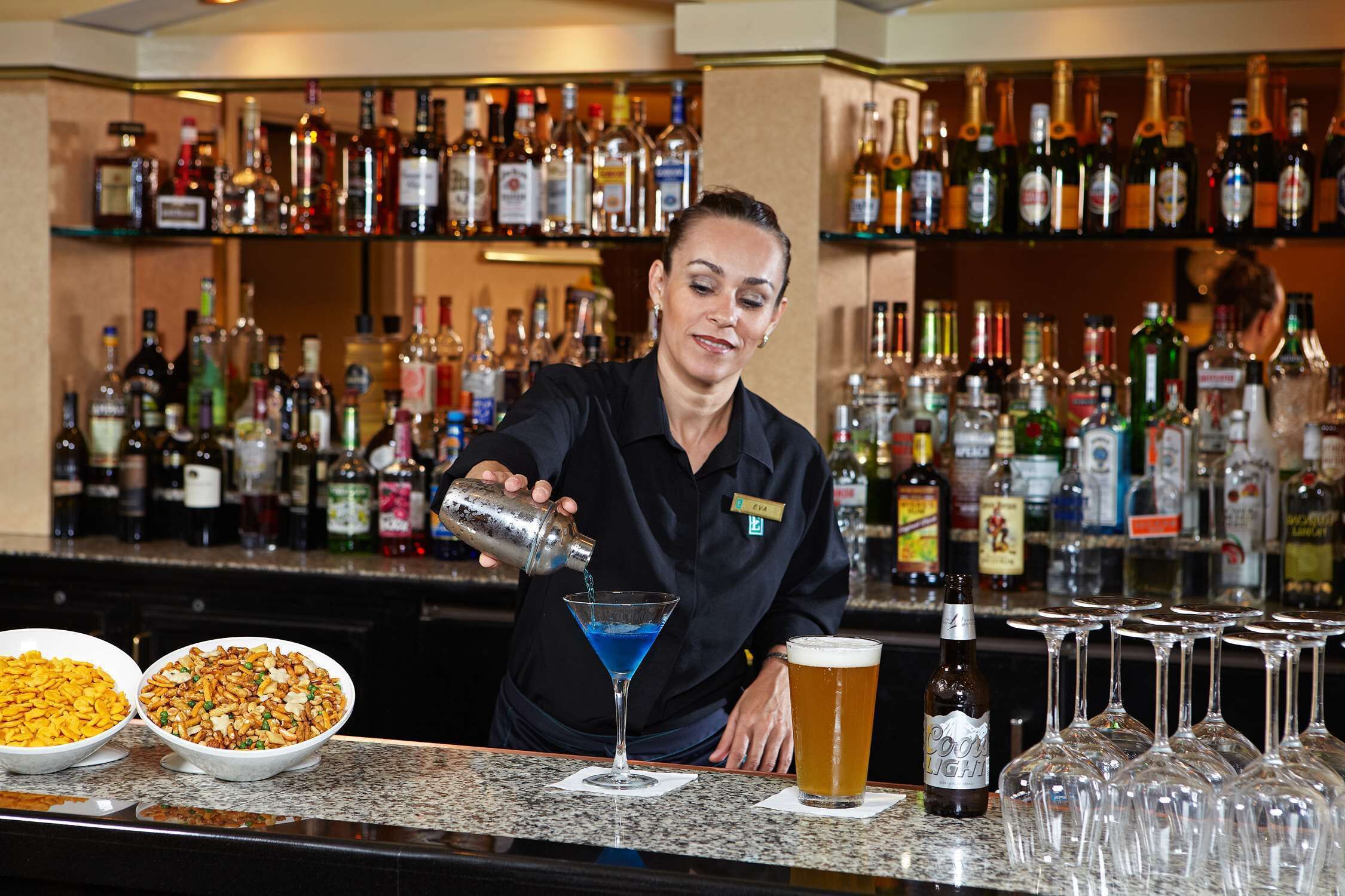 A bartender preparing a cocktail at a bar with various bottles and glasses in the background.