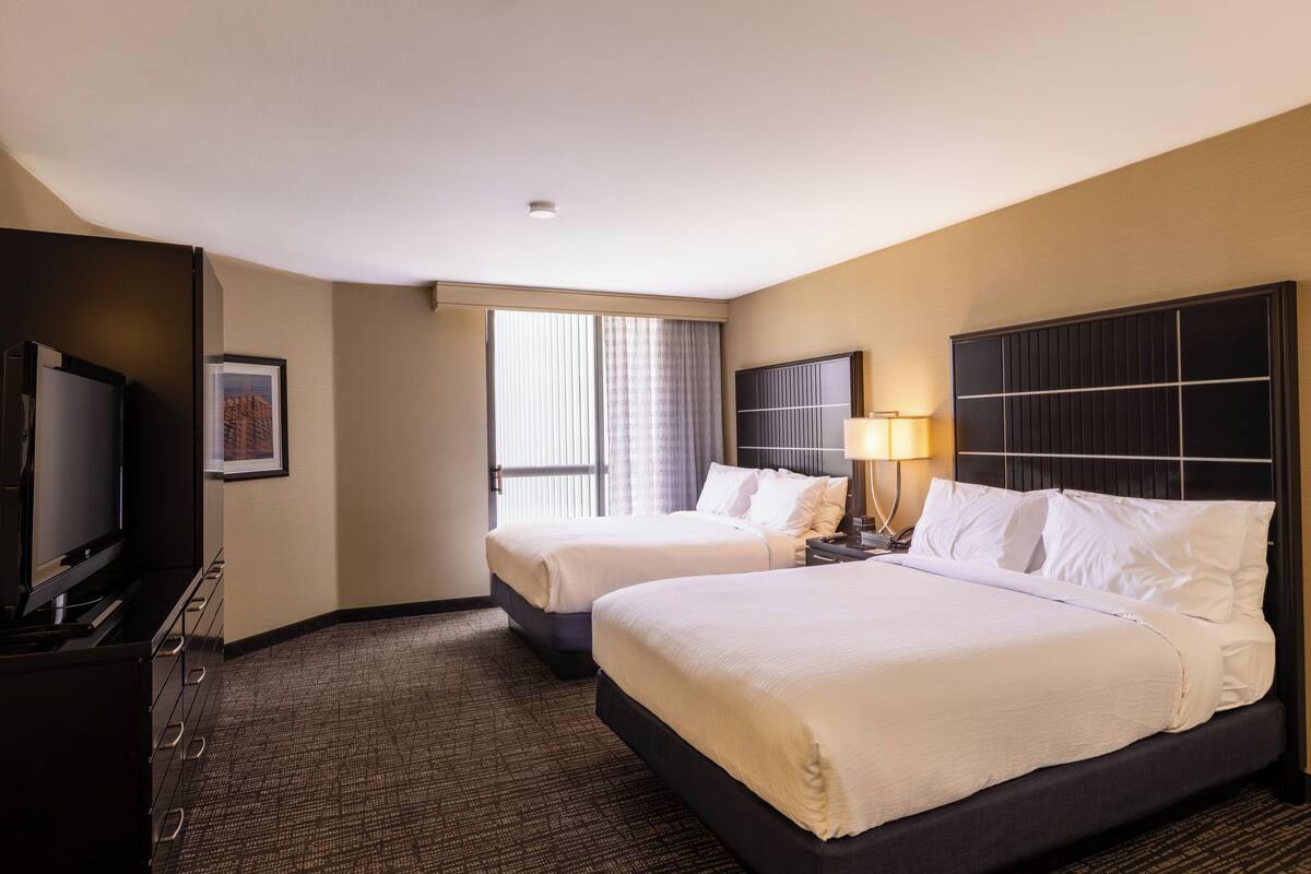 Ein Hotelzimmer mit zwei Betten, einem Fernseher, einem Schreibtisch und einem Fenster mit Vorhängen.