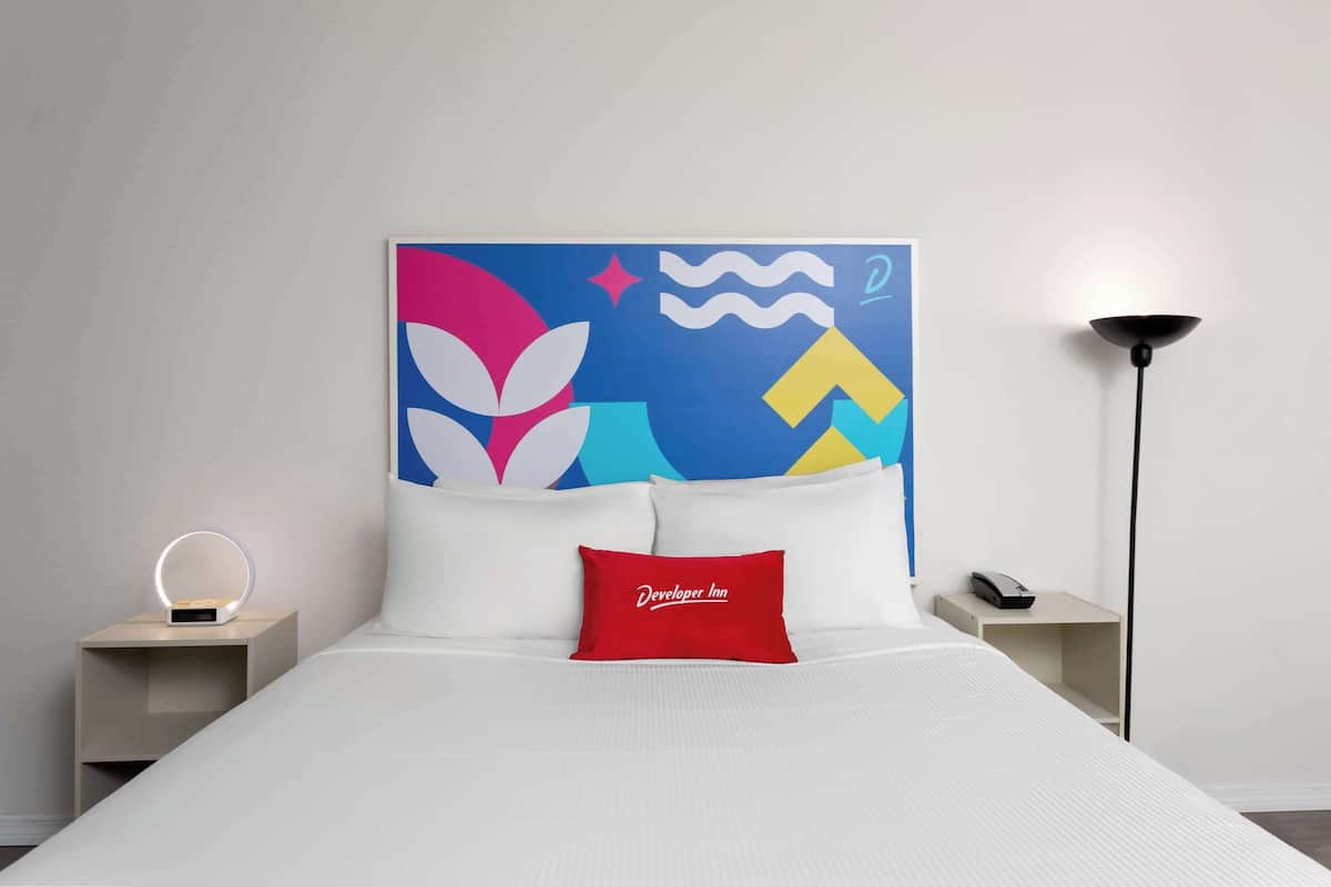 A hotel room with a bed, a red pillow with "Developer Inn" text, a bedside table with a phone, and a wall art piece.