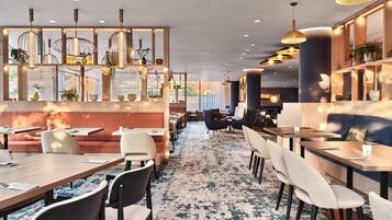 Een modern restaurant met diverse zitmogelijkheden, zoals ruimtes met stoelen en tafels, decoratieve verlichting en een patroonloos tapijt.