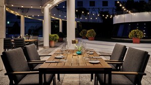An outdoor dining area with a wooden table set for four, surrounded by chairs. There are potted plants and string lights overhead. - Crowne Plaza Bratislava by IHG (Bratislava)
