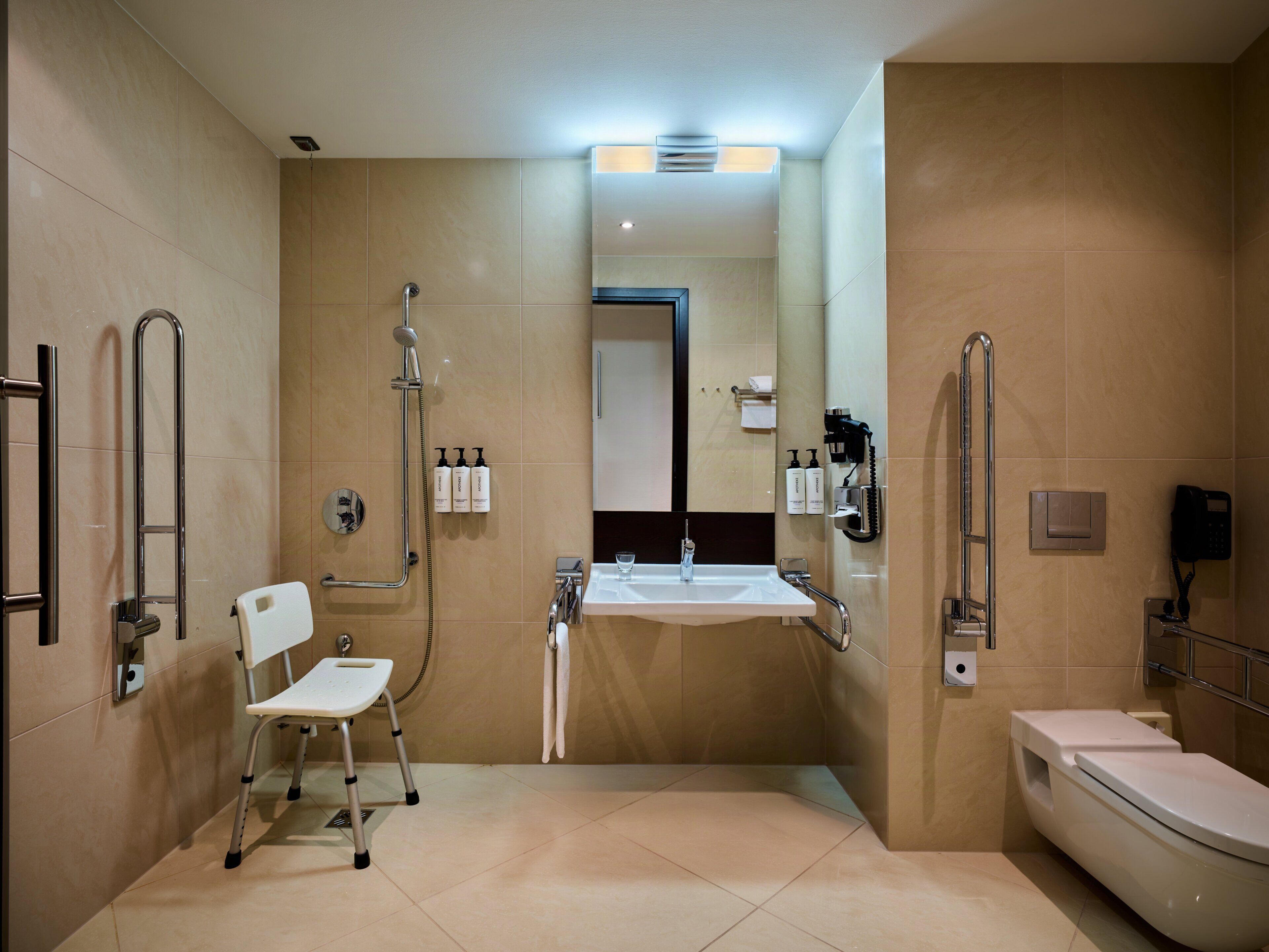 A modern bathroom with a white sink, a toilet, a shower with a handheld showerhead, a white chair, and a handrail.