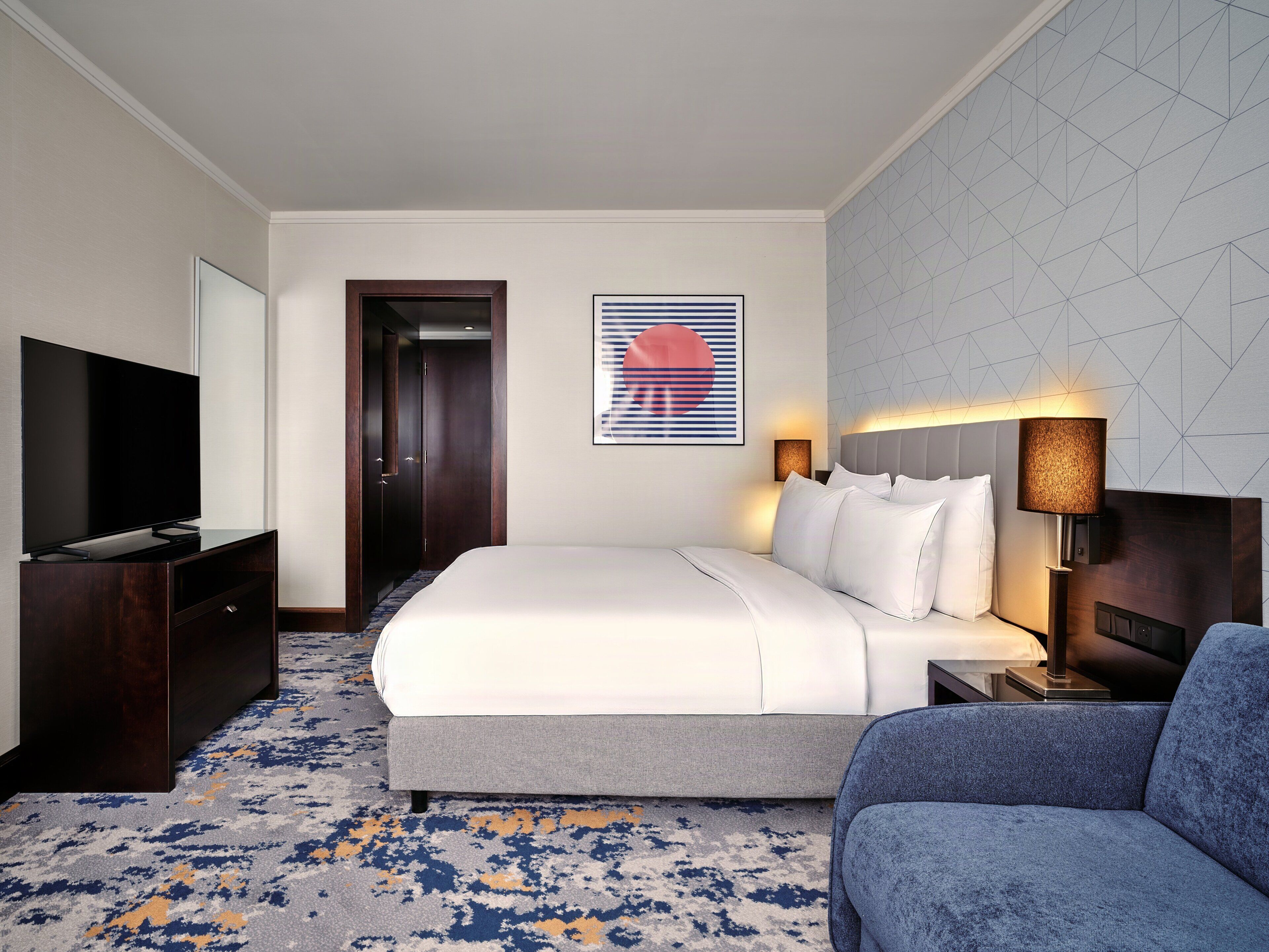 A hotel room with a bed, a TV, a desk, a chair, and a wall with a geometric pattern.