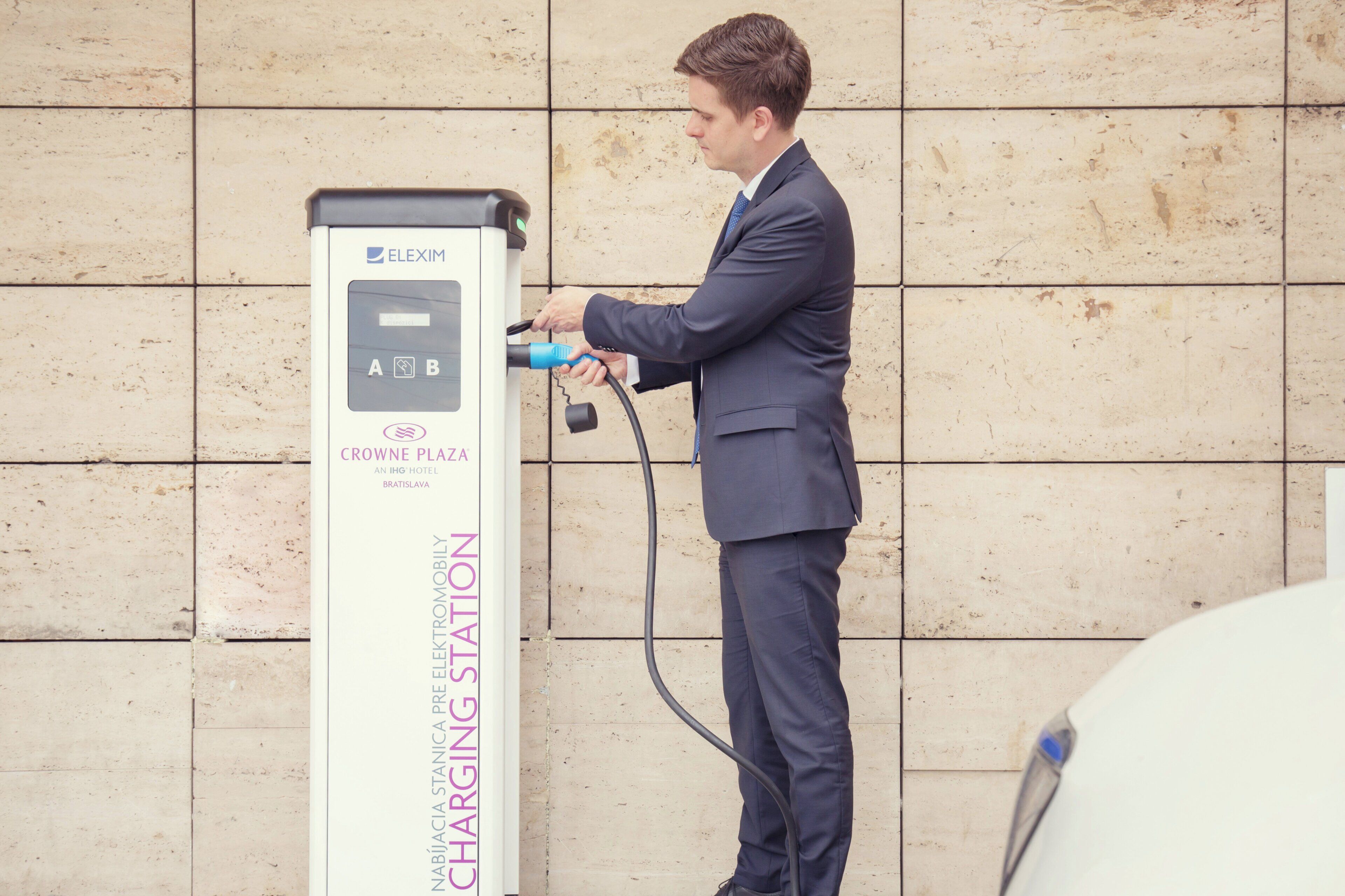 A person charging an electric vehicle at a station labeled 'ELEXIM' with a sign indicating 'NABÍJACIA CHARGING STATION.'
