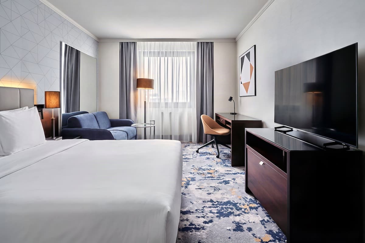 A hotel room with a bed, a desk, a chair, a TV, a lamp, and a window with curtains.