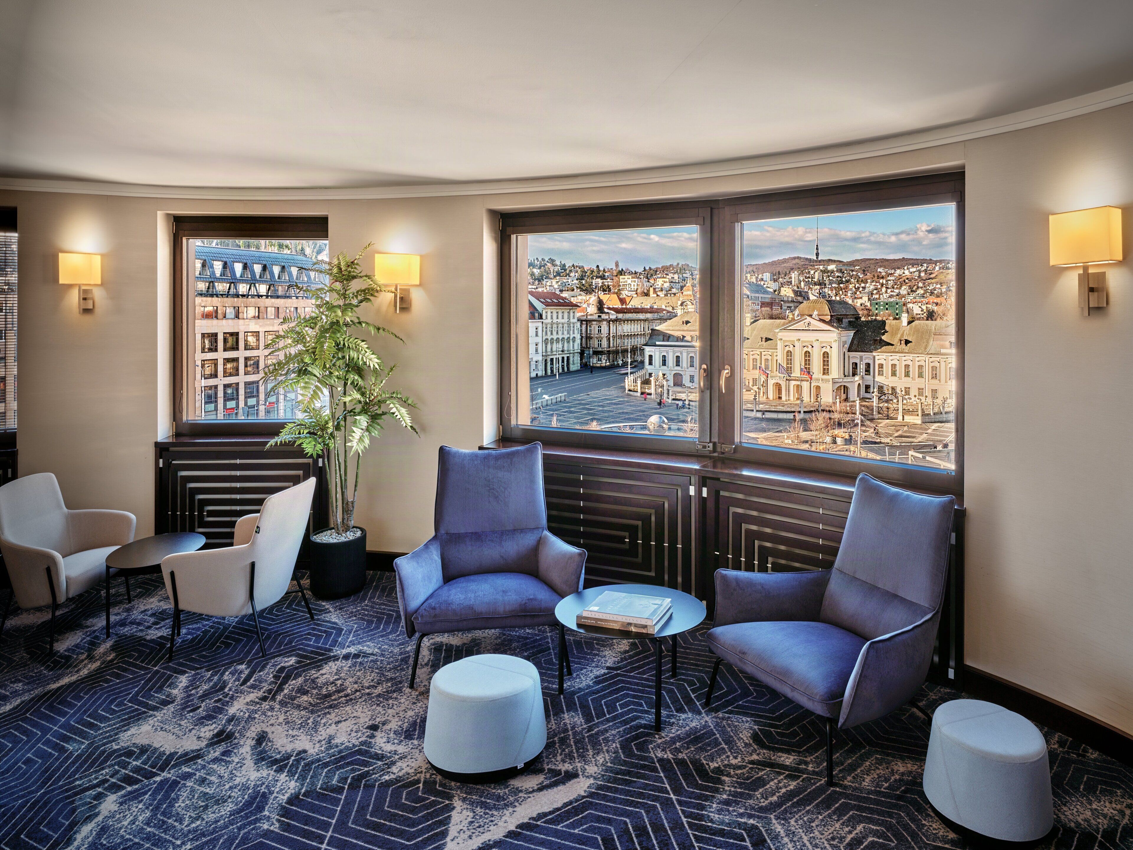 Ein Hotelzimmer mit einer gebogenen Sitzecke, einer Topfpflanze und großen Fenstern mit Blick auf die Stadt.