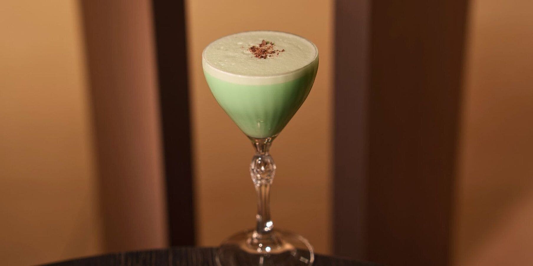 A cocktail in a martini glass with a green rim, placed on a table.