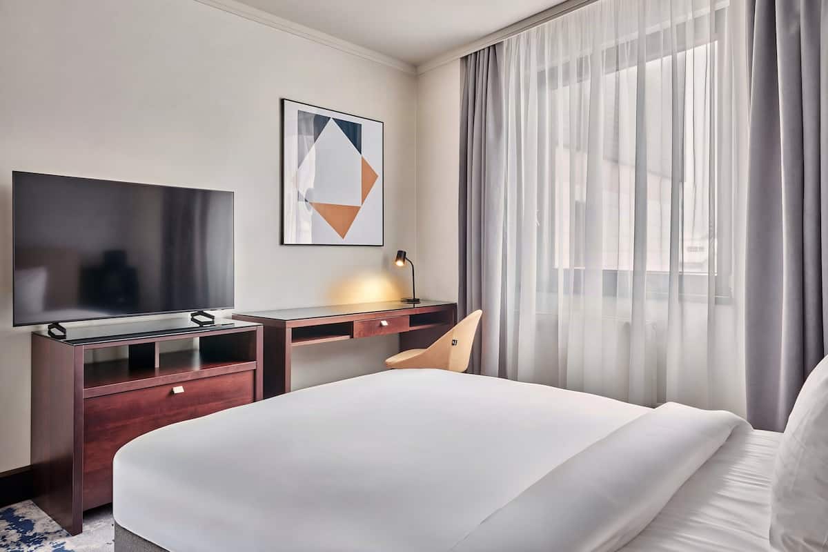 A hotel room with a bed, a desk with a computer, a TV, and a window with curtains.