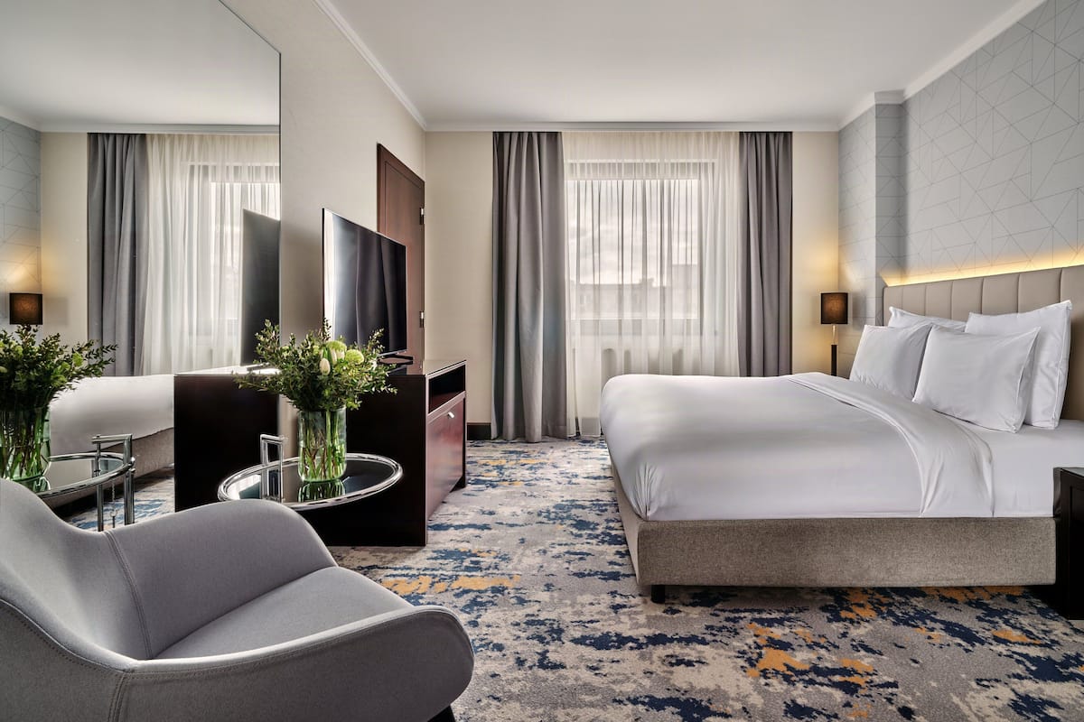 A hotel room with a bed, a chair, a TV, a desk, and a vase with flowers.