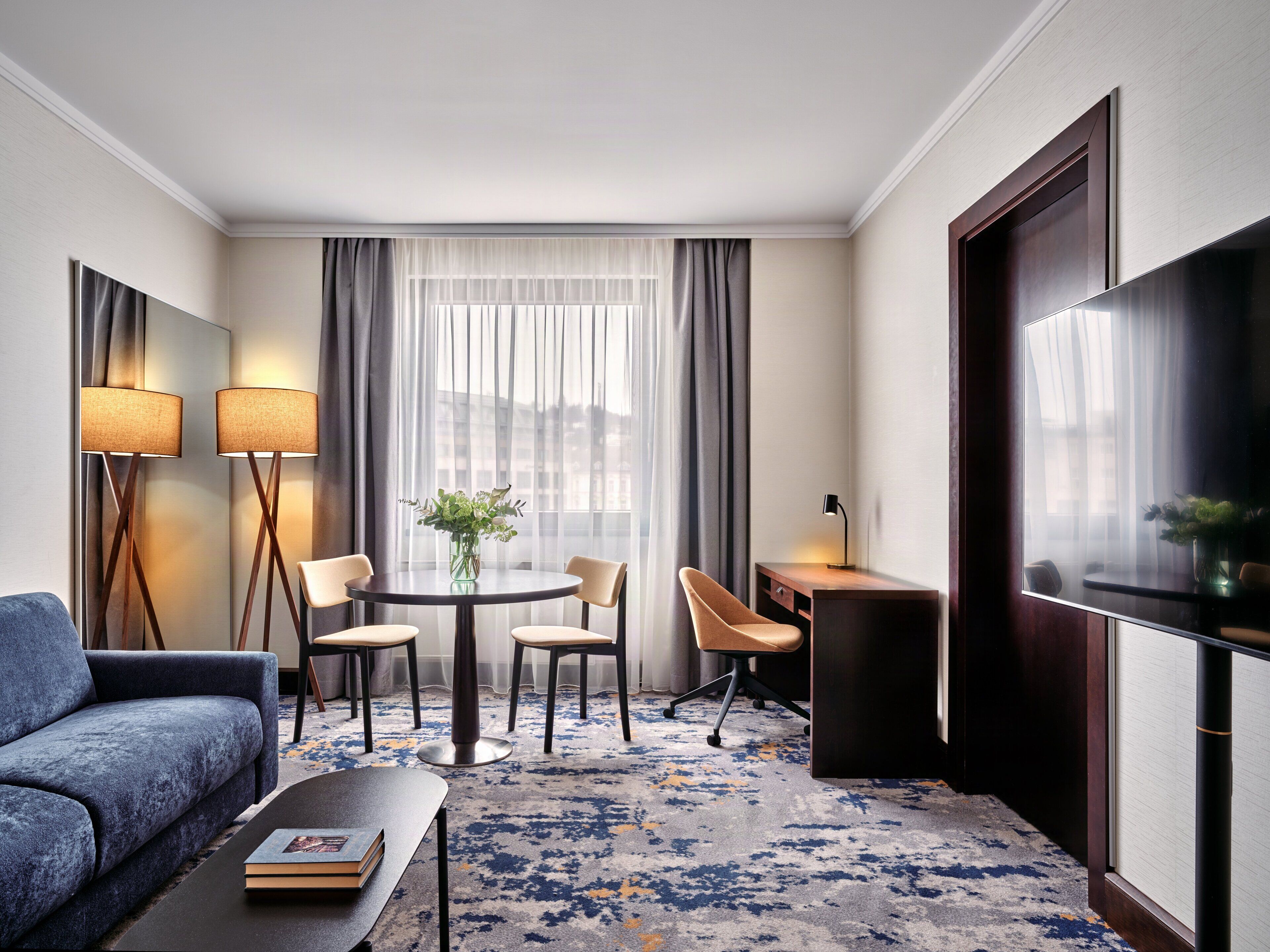 A hotel room with a sofa, a small round table with chairs, a desk, a TV, and a large window with curtains.