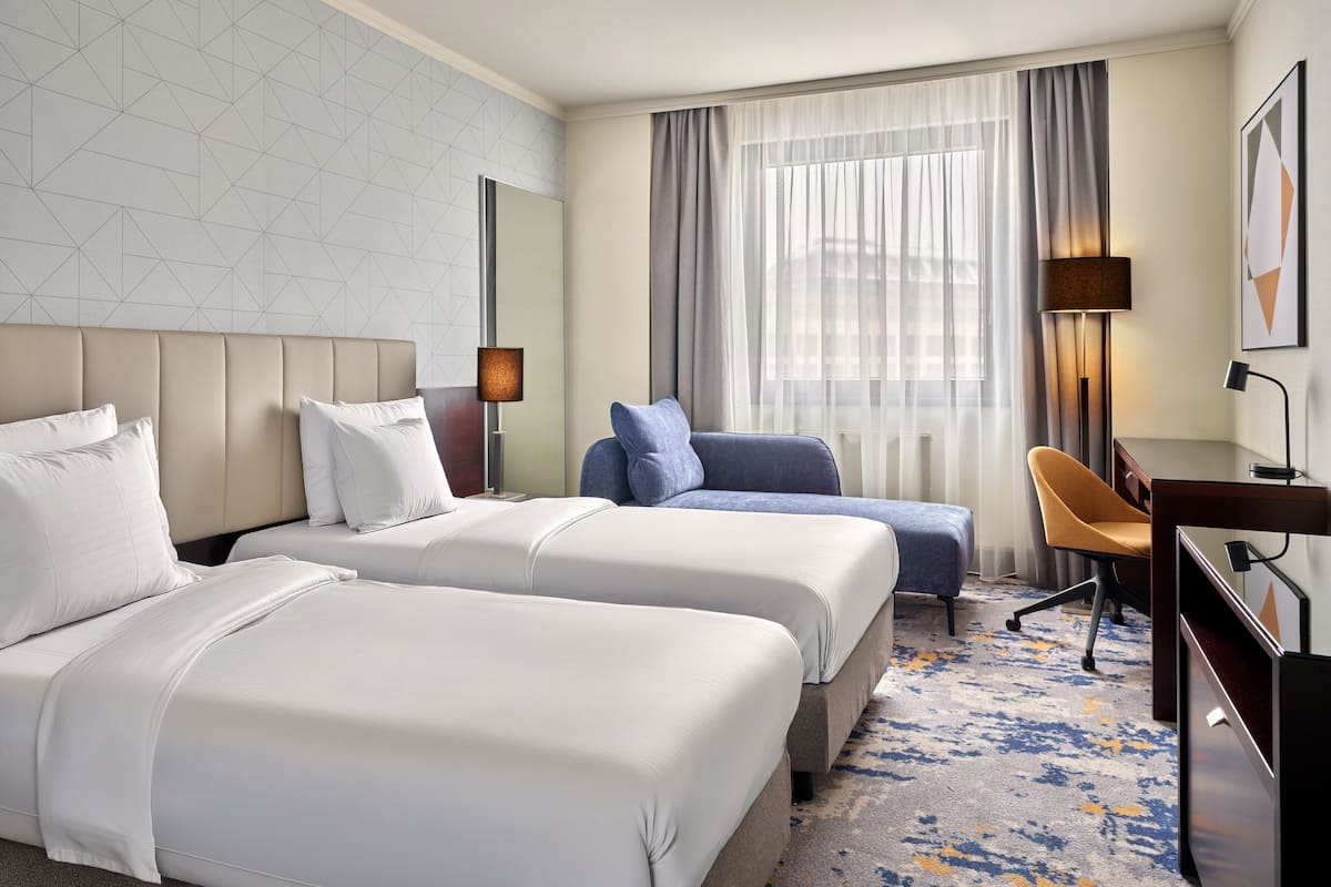 A hotel room with two beds, a desk, a chair, a lamp, and a window with curtains.